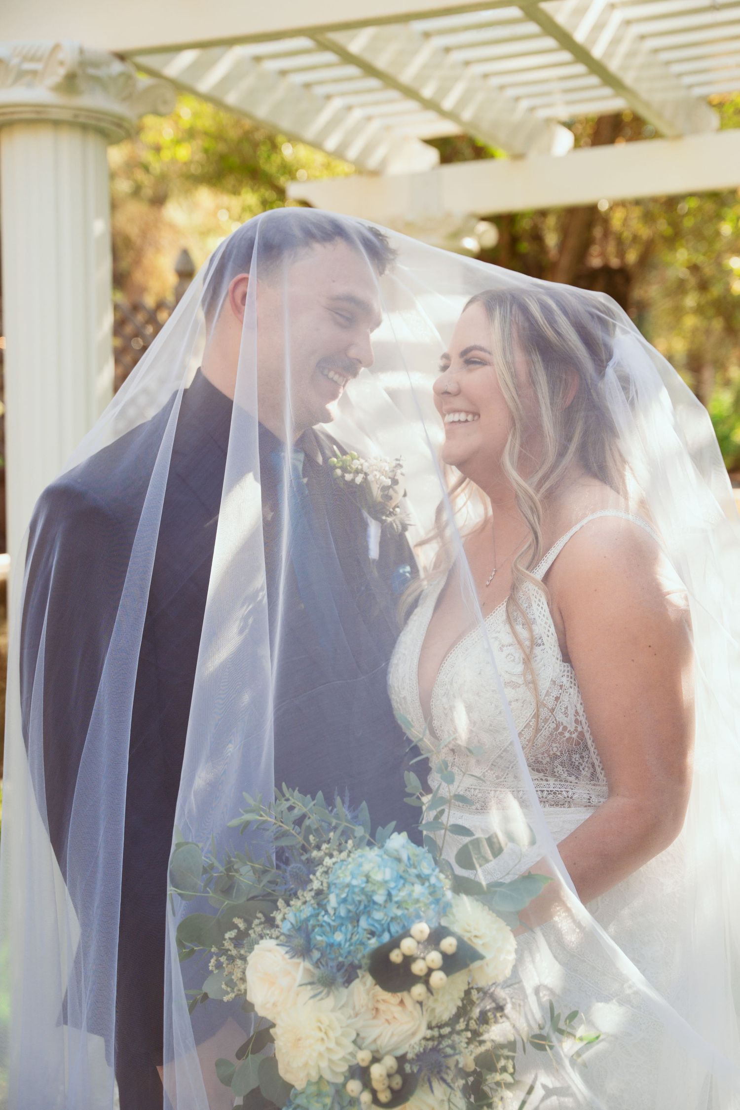 A romantic wedding couple shares an intimate moment under a flowing veil in golden sunlight.