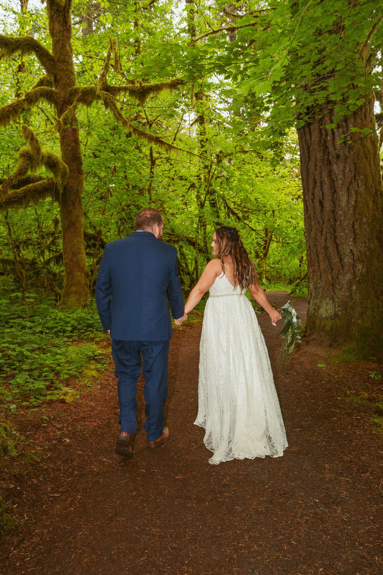 A couple in wedding attire walking hand in hand down a forest path surrounded by green trees.