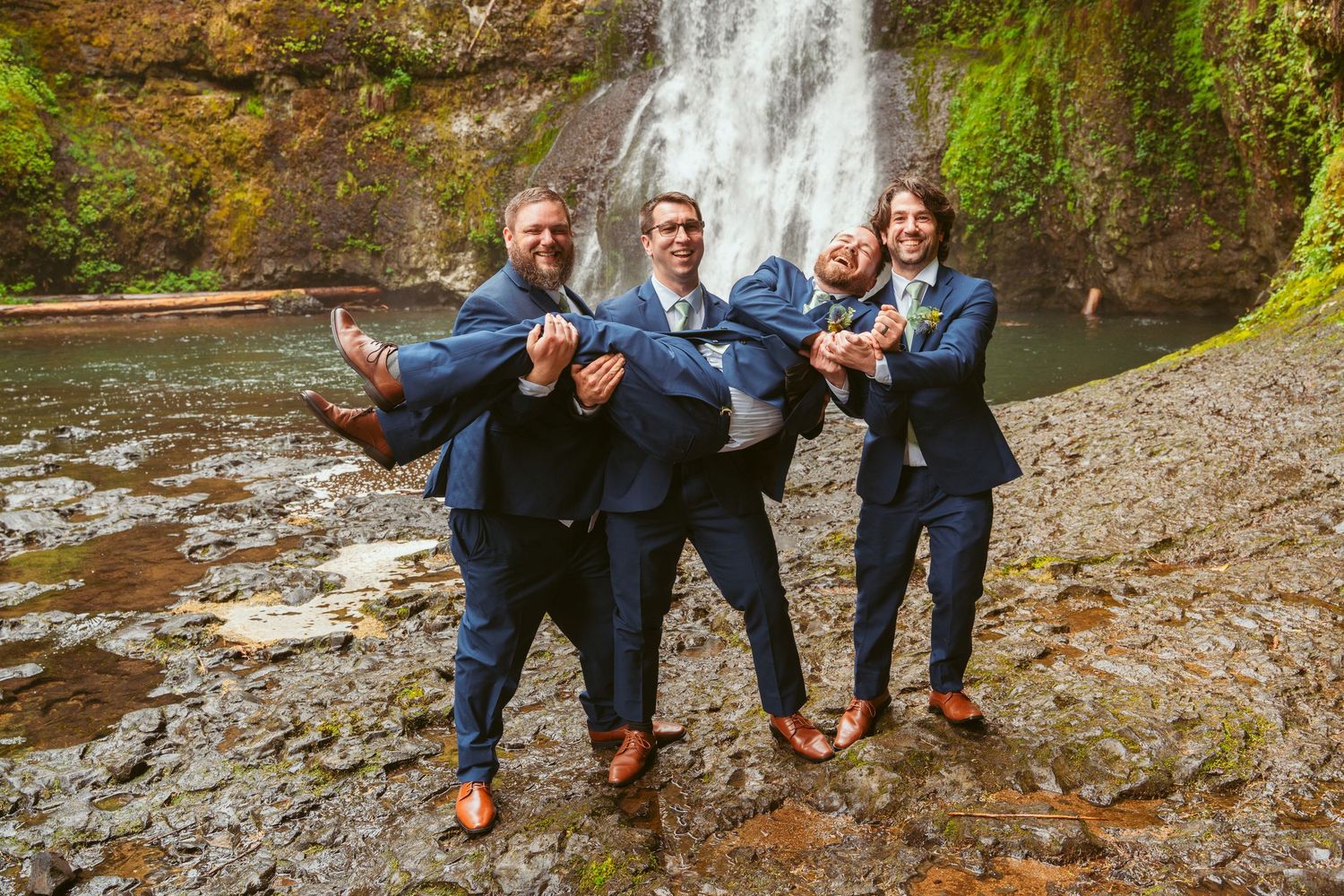 Groomsmen strike fun poses near a waterfall wearing matching navy suits.