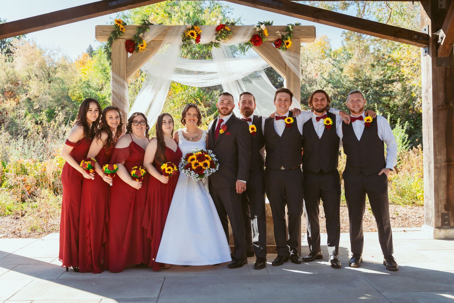 Full wedding party lined up under rustic outdoor pavilion with red and black attire.