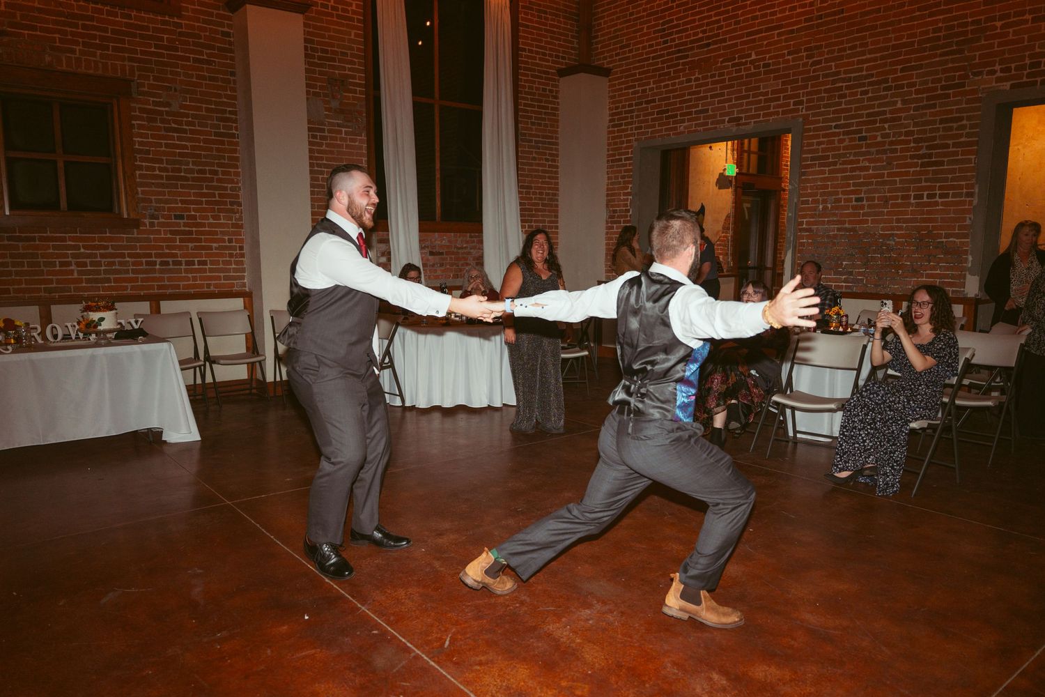 Two people dancing energetically on a wooden dance floor in front of exposed brick walls at a reception venue.