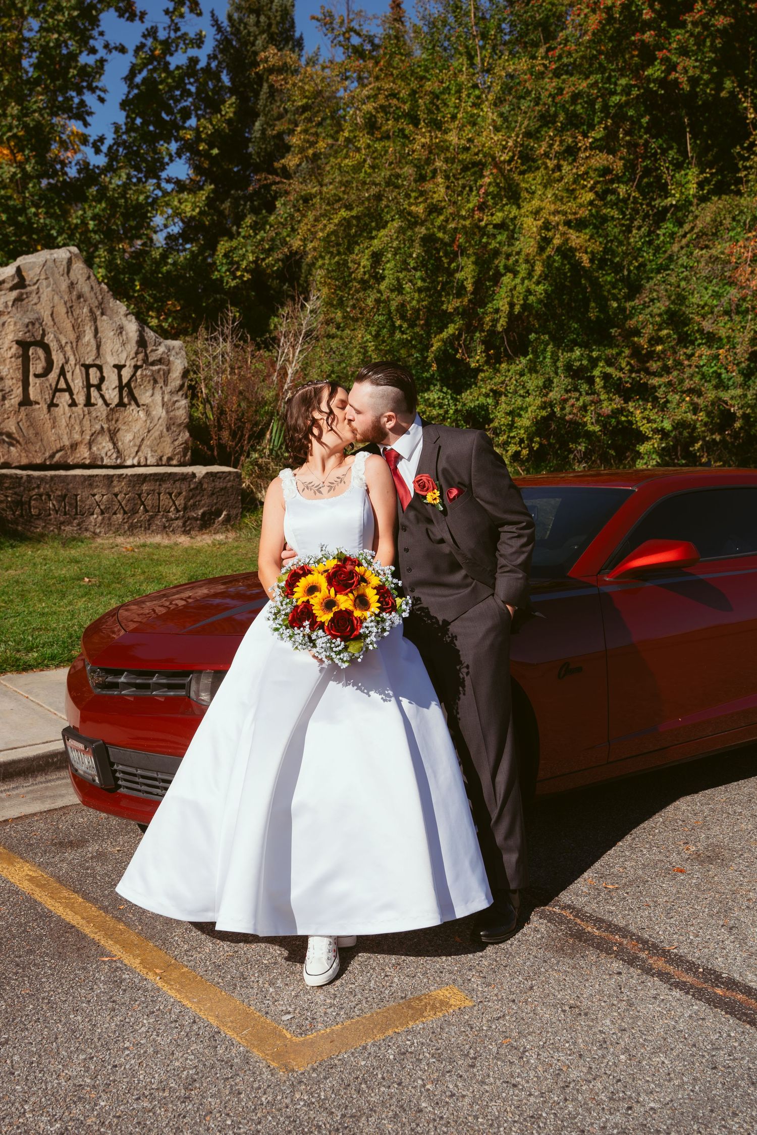 Newlyweds pose with sunflower bouquet next to red classic car in autumn setting.