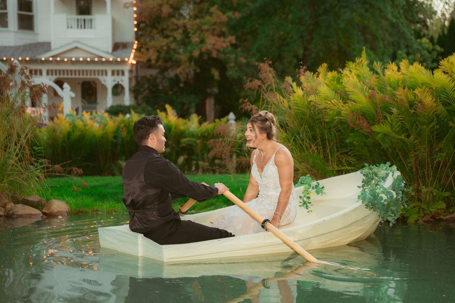 Wedding couple sitting in rowboat on pond near Victorian-style house.
