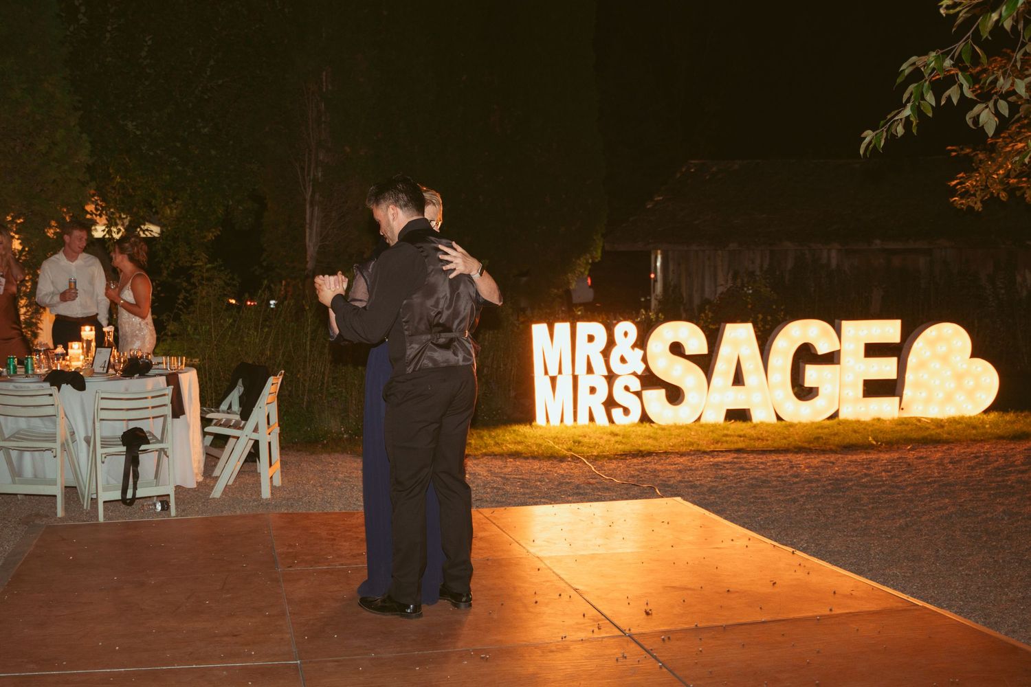 Couple dancing near illuminated Mr & Mrs Sage sign at nighttime wedding reception.