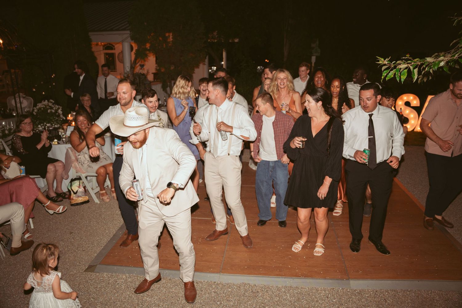 Large group of wedding guests dancing together at an outdoor reception.