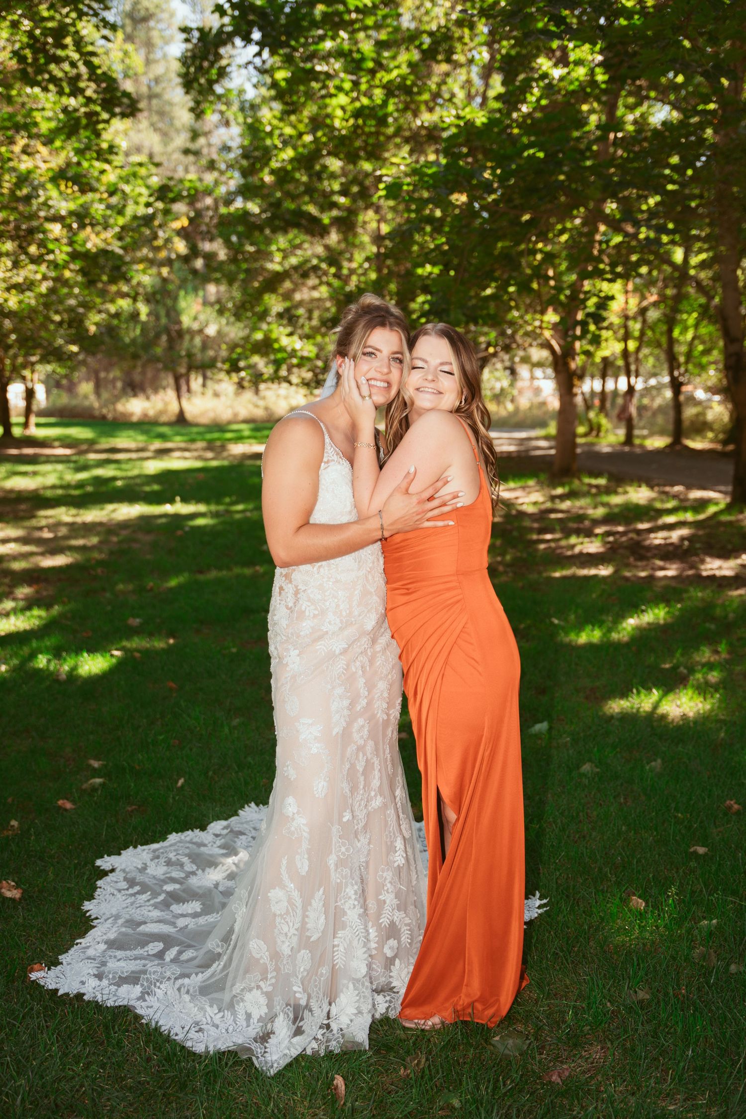 Two people in formal attire, one wearing white lace and another in orange, share a joyful moment in a sunny garden setting.