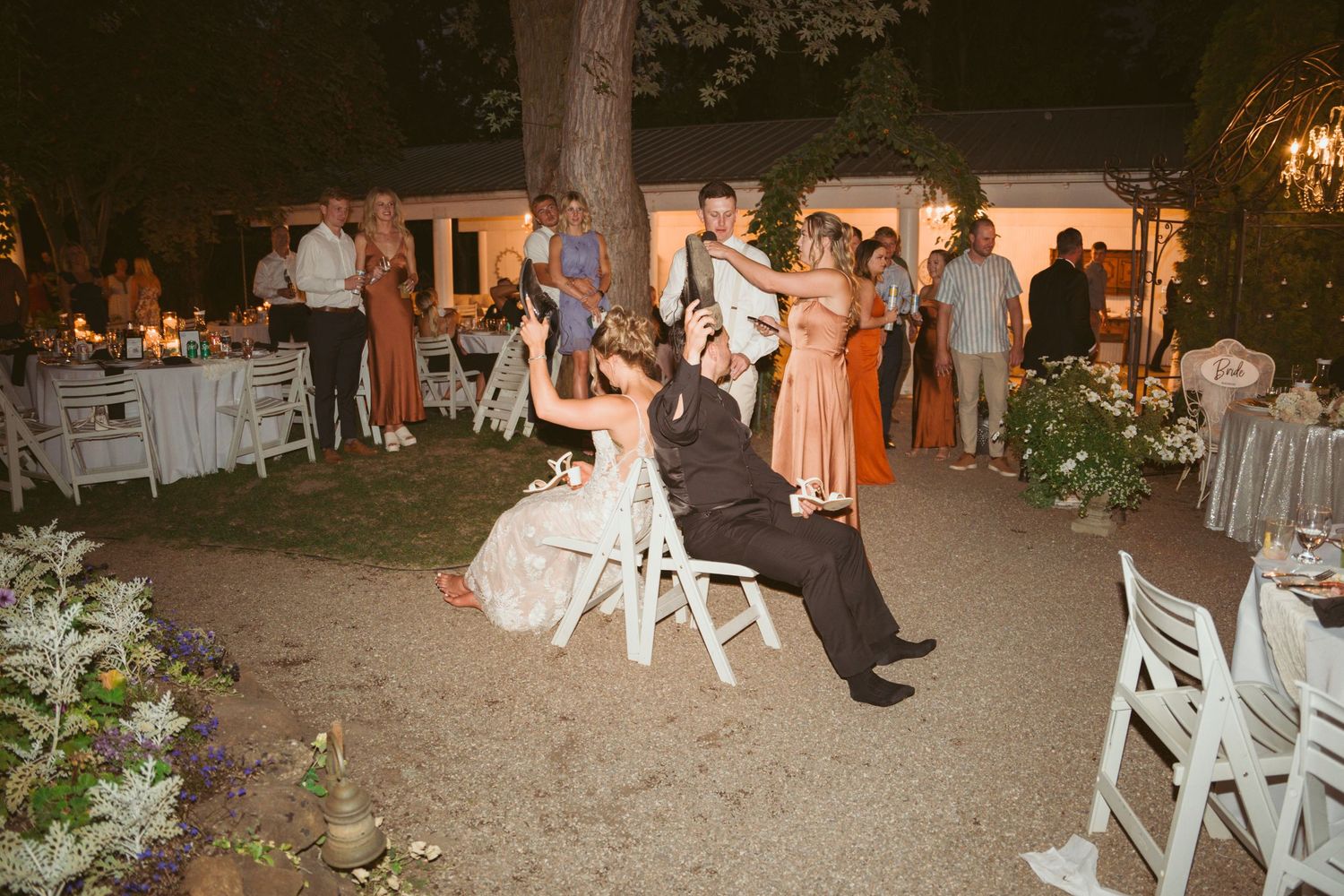 Outdoor wedding reception scene with guests mingling under trees and string lights at dusk.