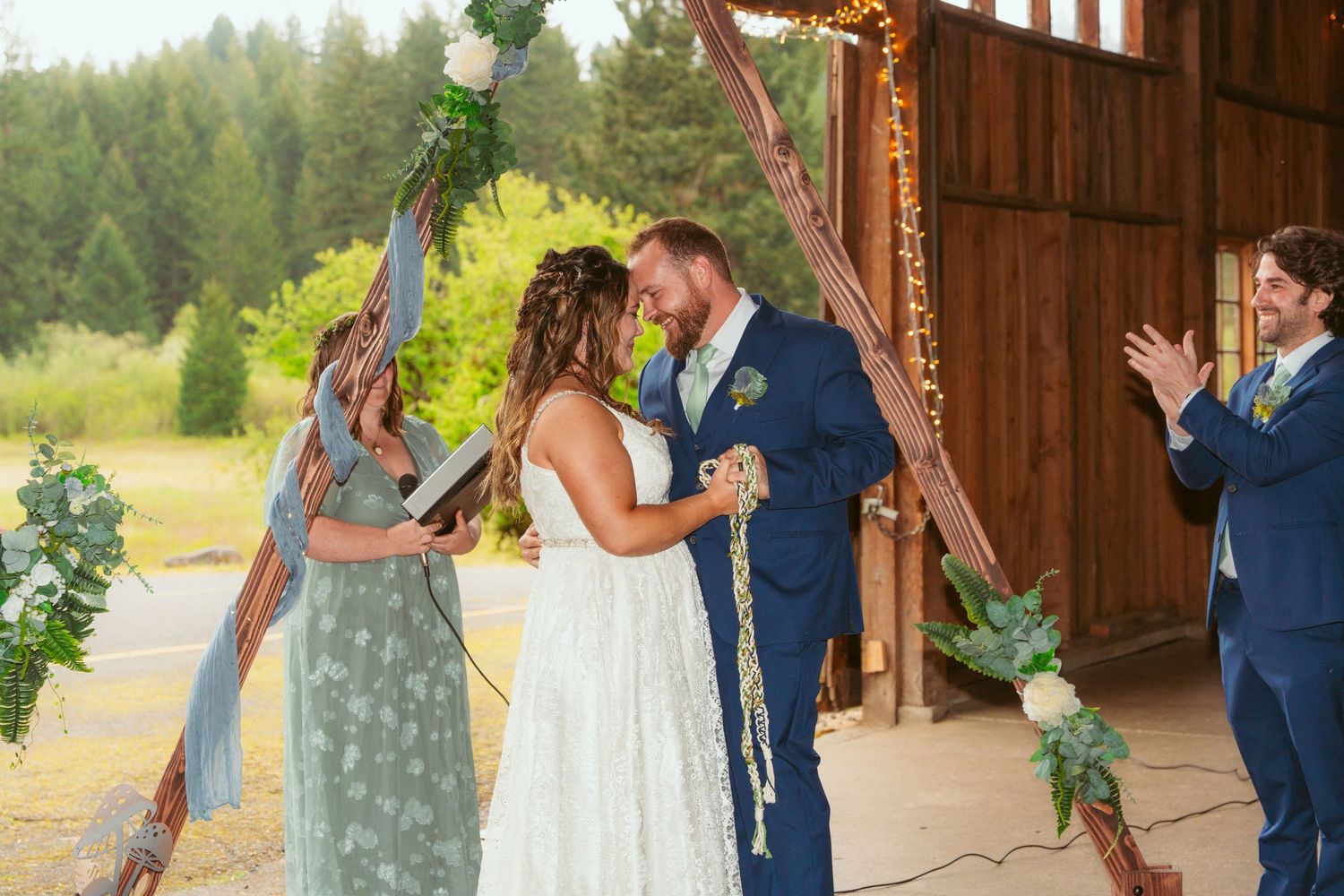 Wedding ceremony taking place in a rustic barn with forest views through open walls.