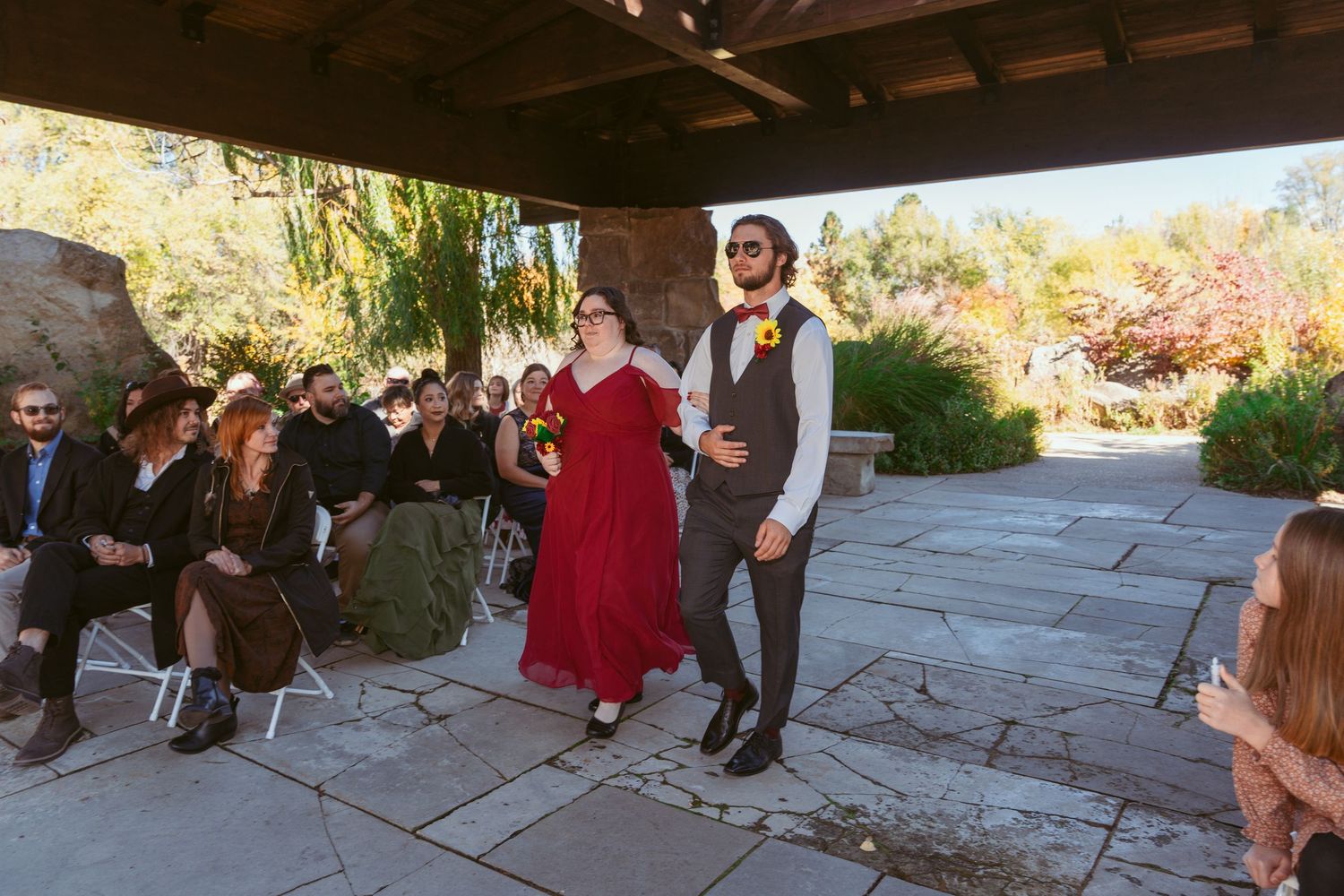 A group of people gather under a covered patio area surrounded by autumn foliage.