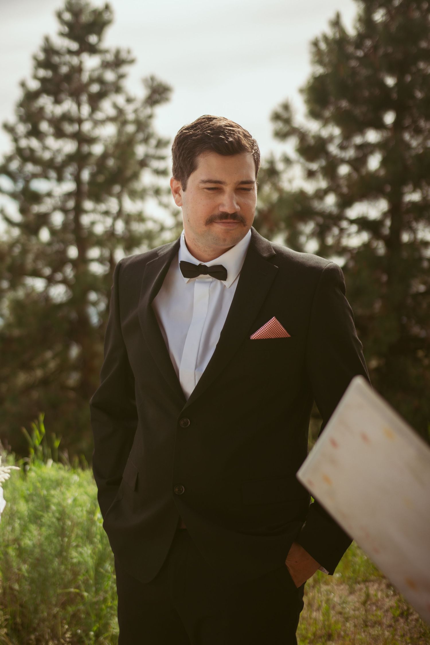 A groom in a black tuxedo and bowtie smiles during an outdoor mountain ceremony.