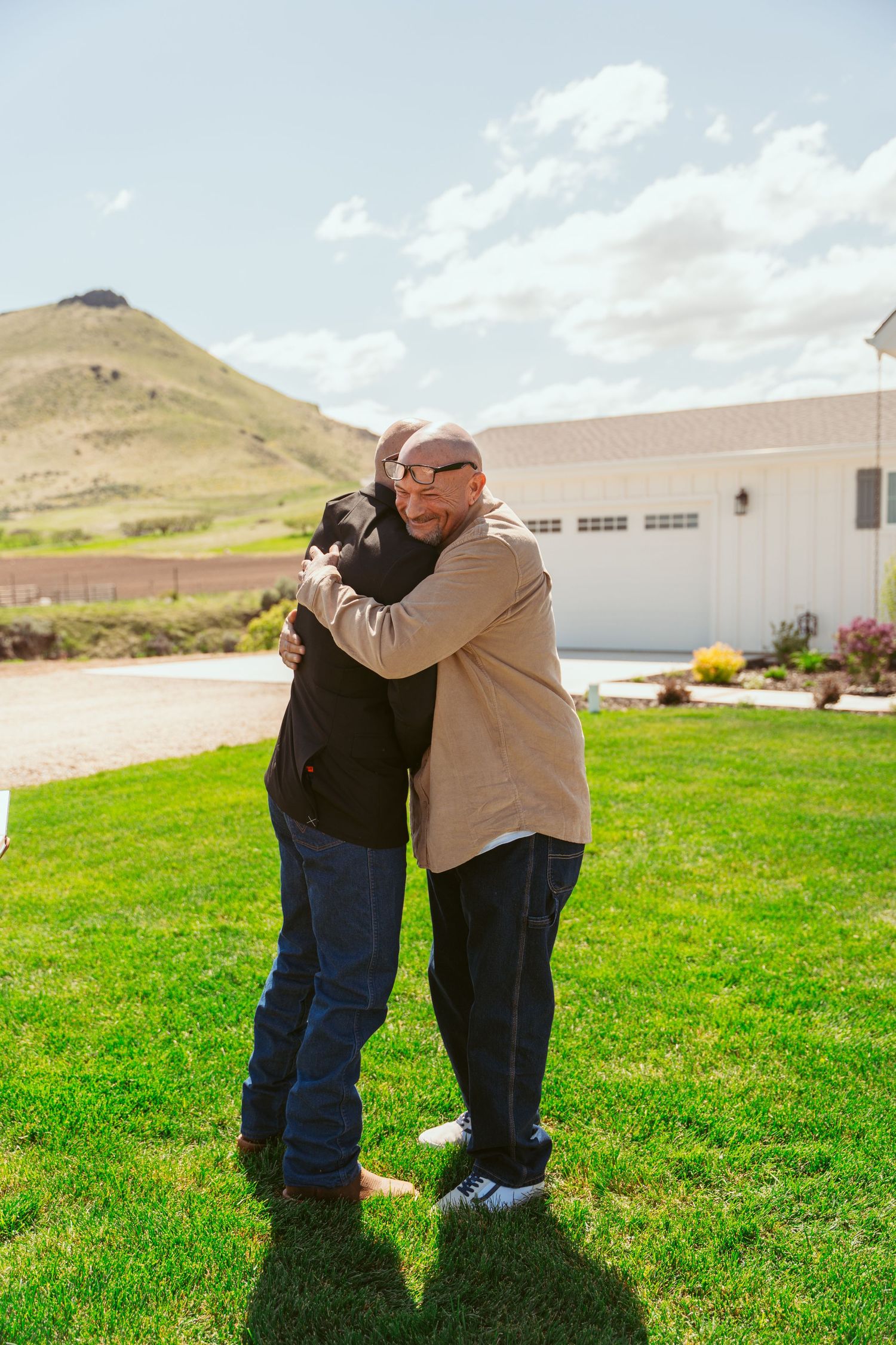 Emotional embrace on sunny lawn with mountains and white buildings in background.