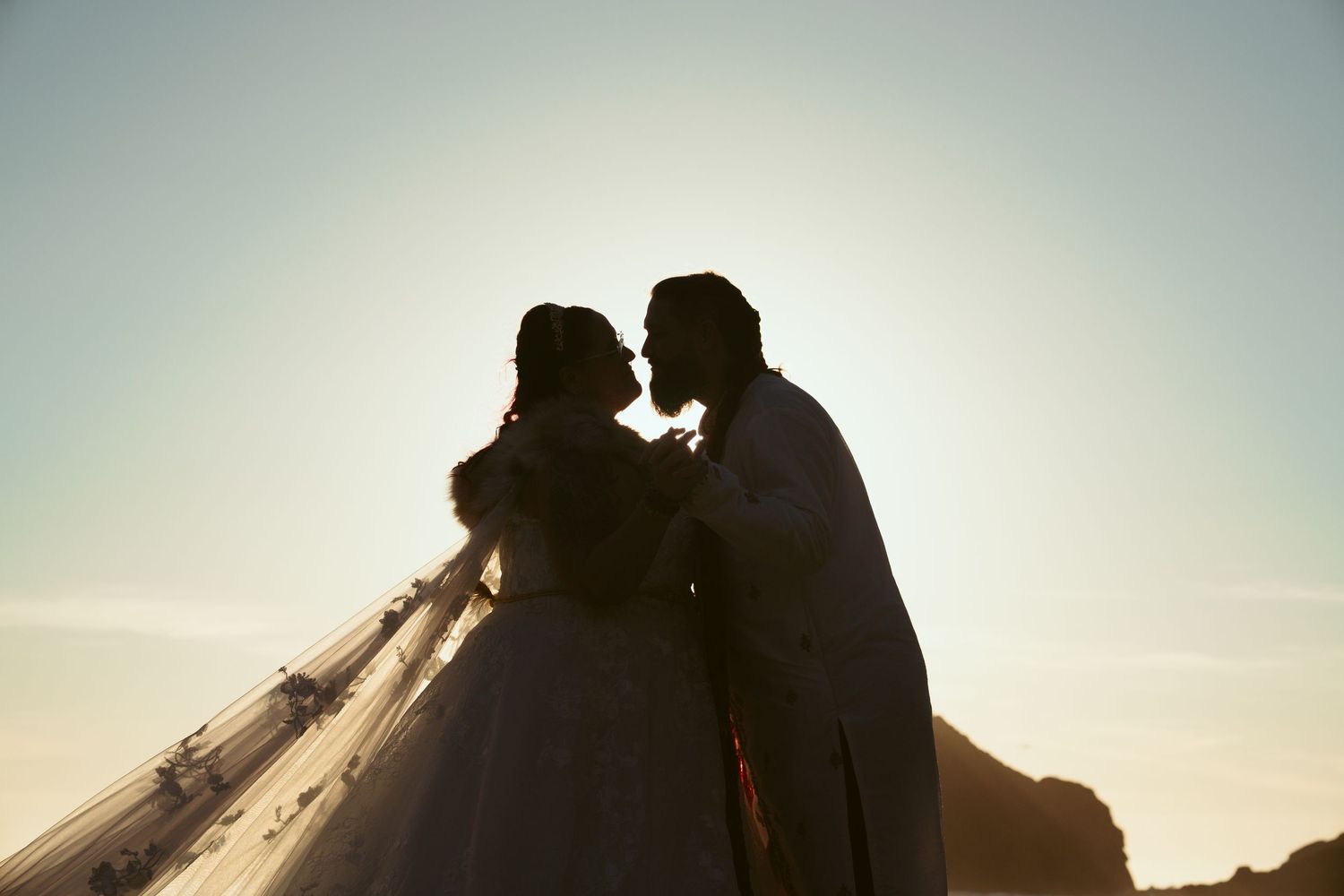Silhouetted couple in wedding attire share a tender moment against the setting sun.
