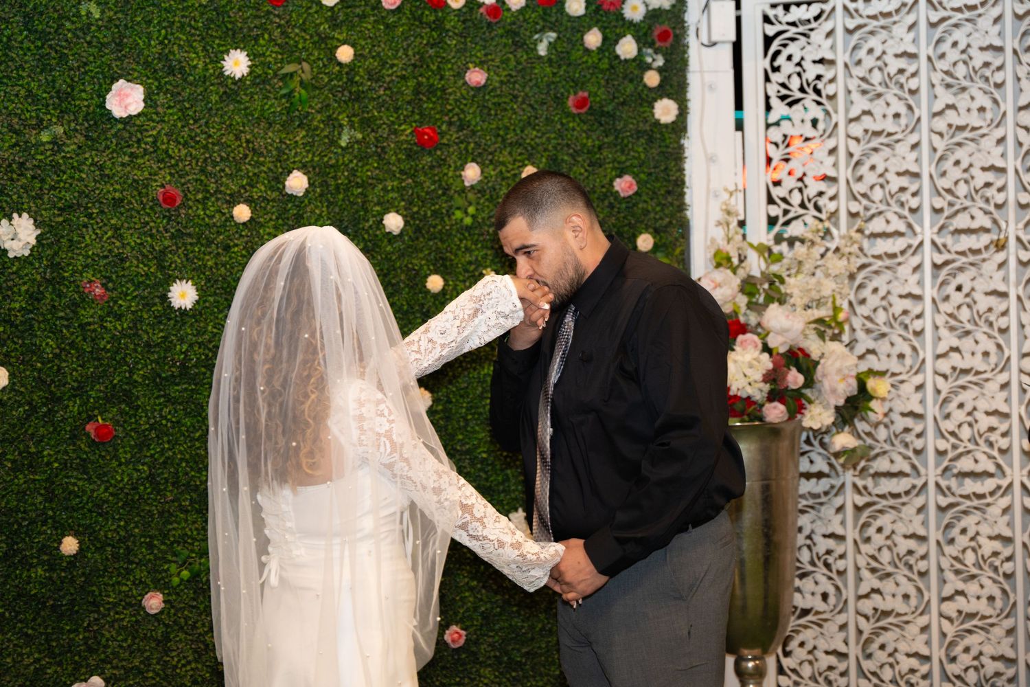 Newlyweds share a moment in front of a floral wall with string lights.