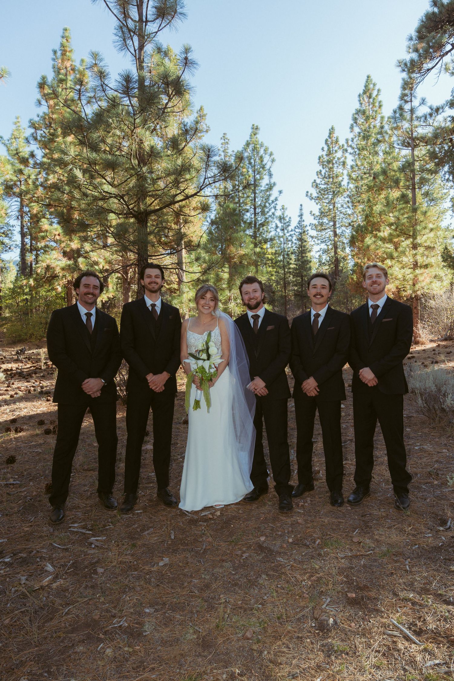 Wedding party group photo with groomsmen in black suits and bridesmaids in forest setting.