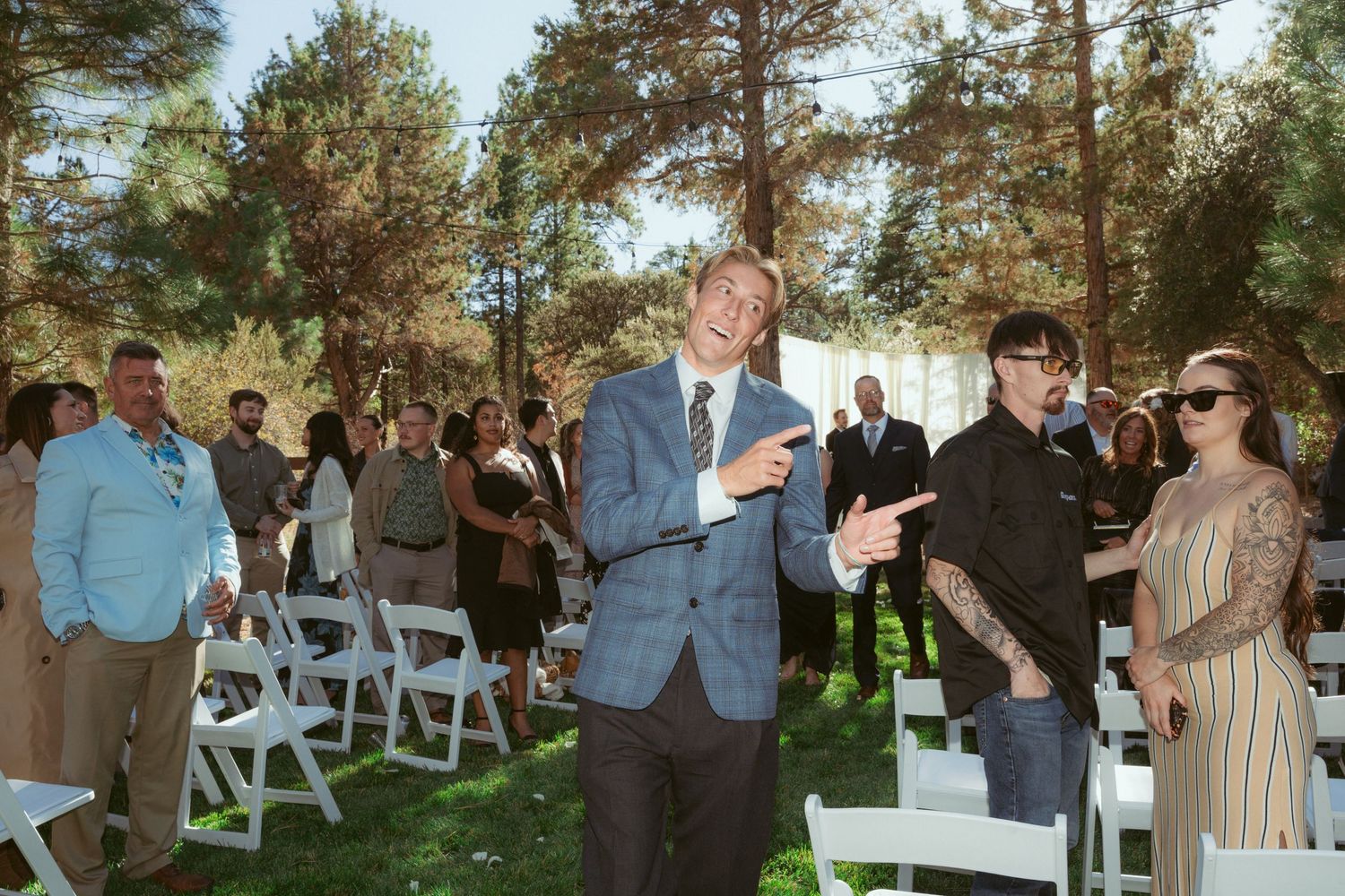 Outdoor wedding celebration with guests mingling among white chairs under tall pines.