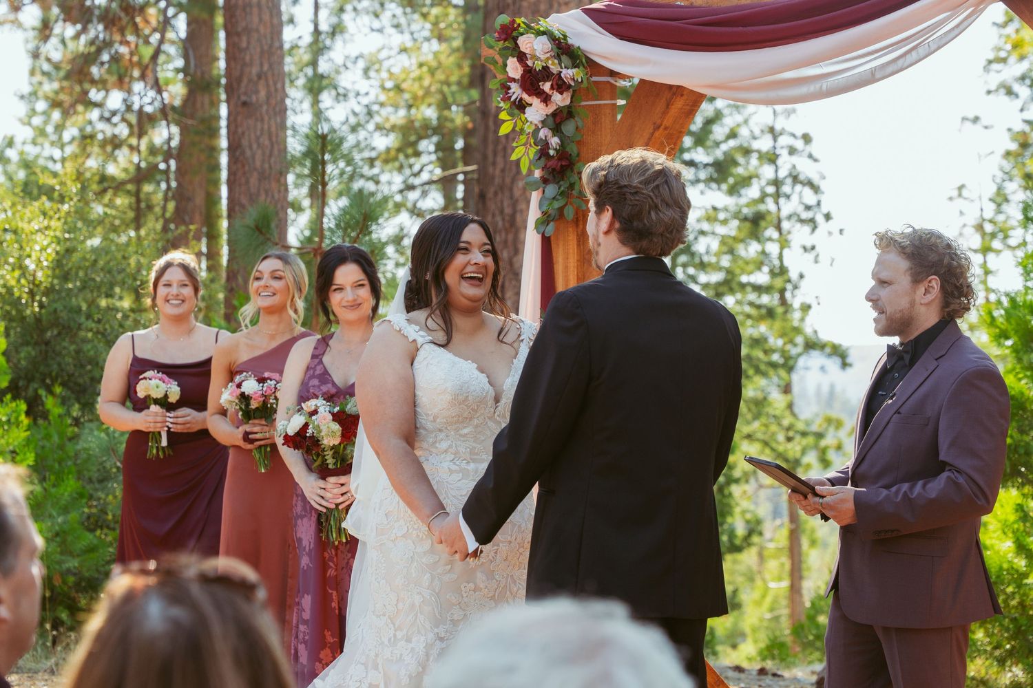 Forest wedding ceremony with burgundy draped arch and pine trees in background.