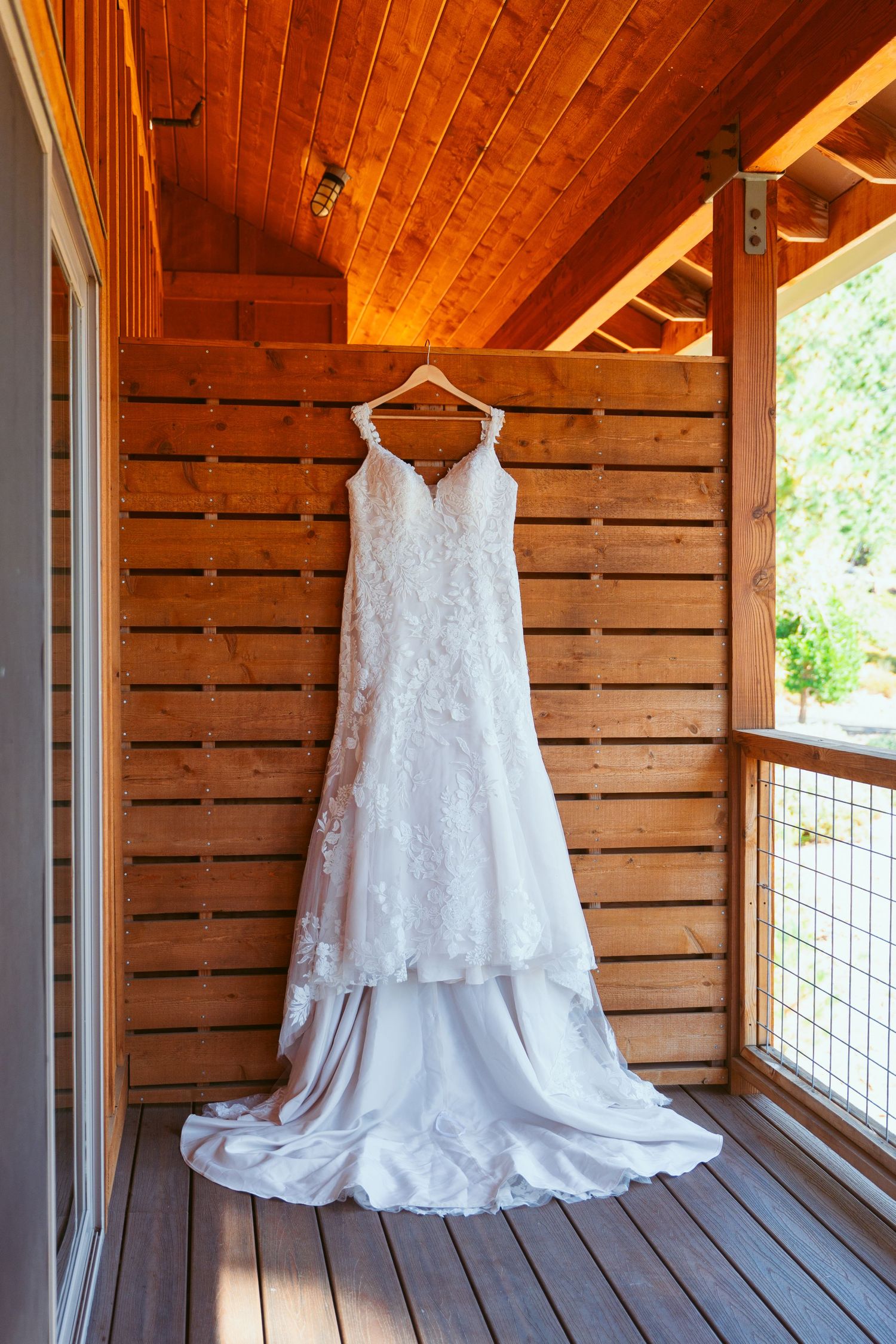White lace wedding dress hanging on wooden porch with natural lighting.