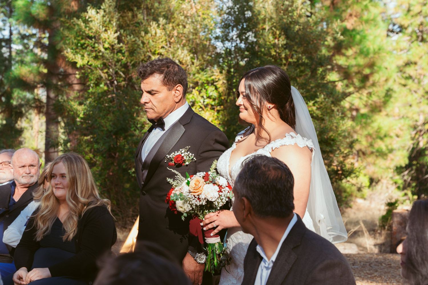 Wedding ceremony procession in a sunlit forest setting with guests seated.