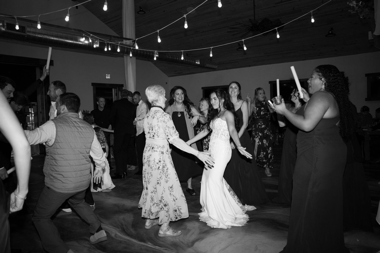 Black and white photo of wedding guests dancing at an indoor reception with string lights.