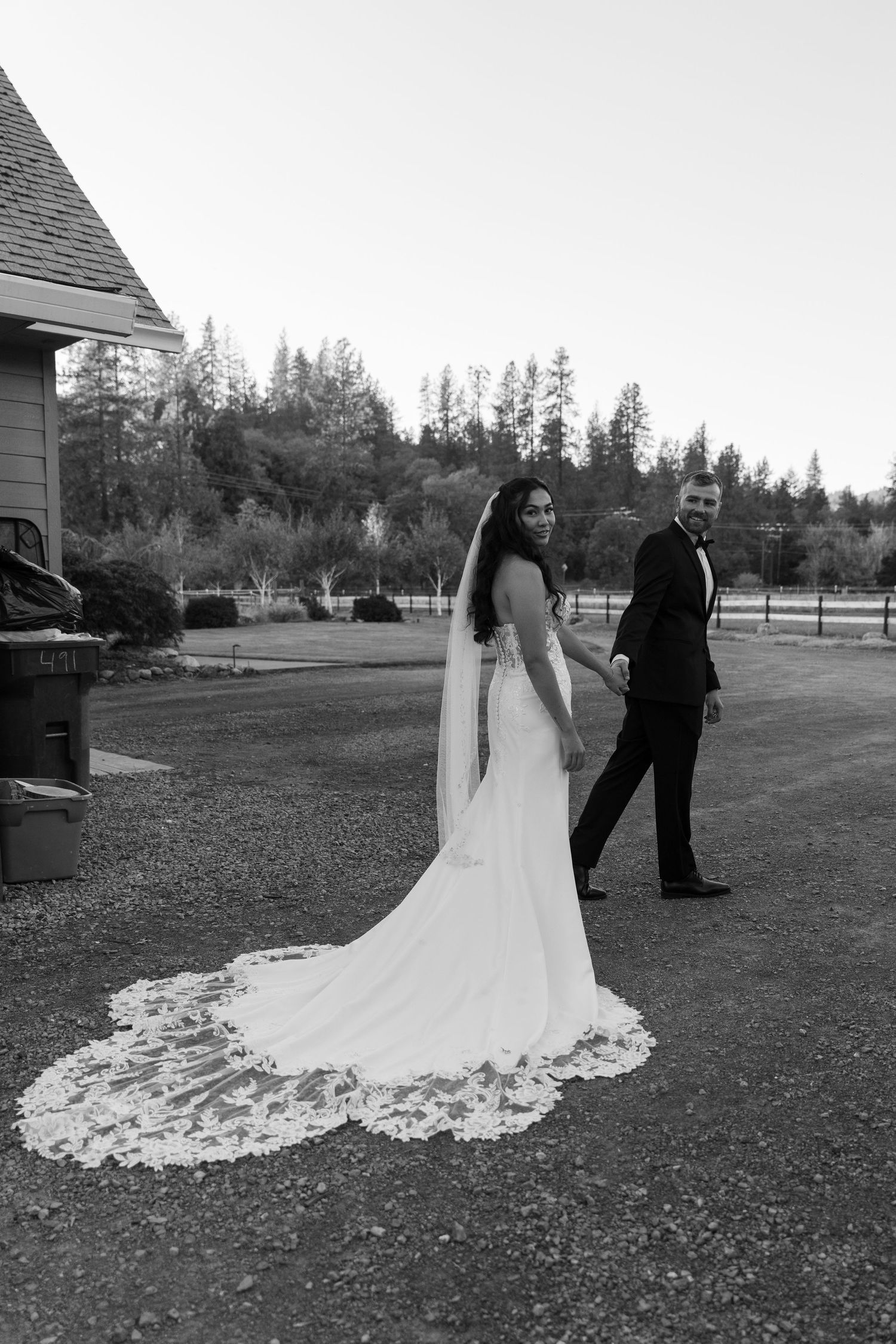 A wedding couple poses on a grassy lawn with a stunning long lace train on the bride's white gown.