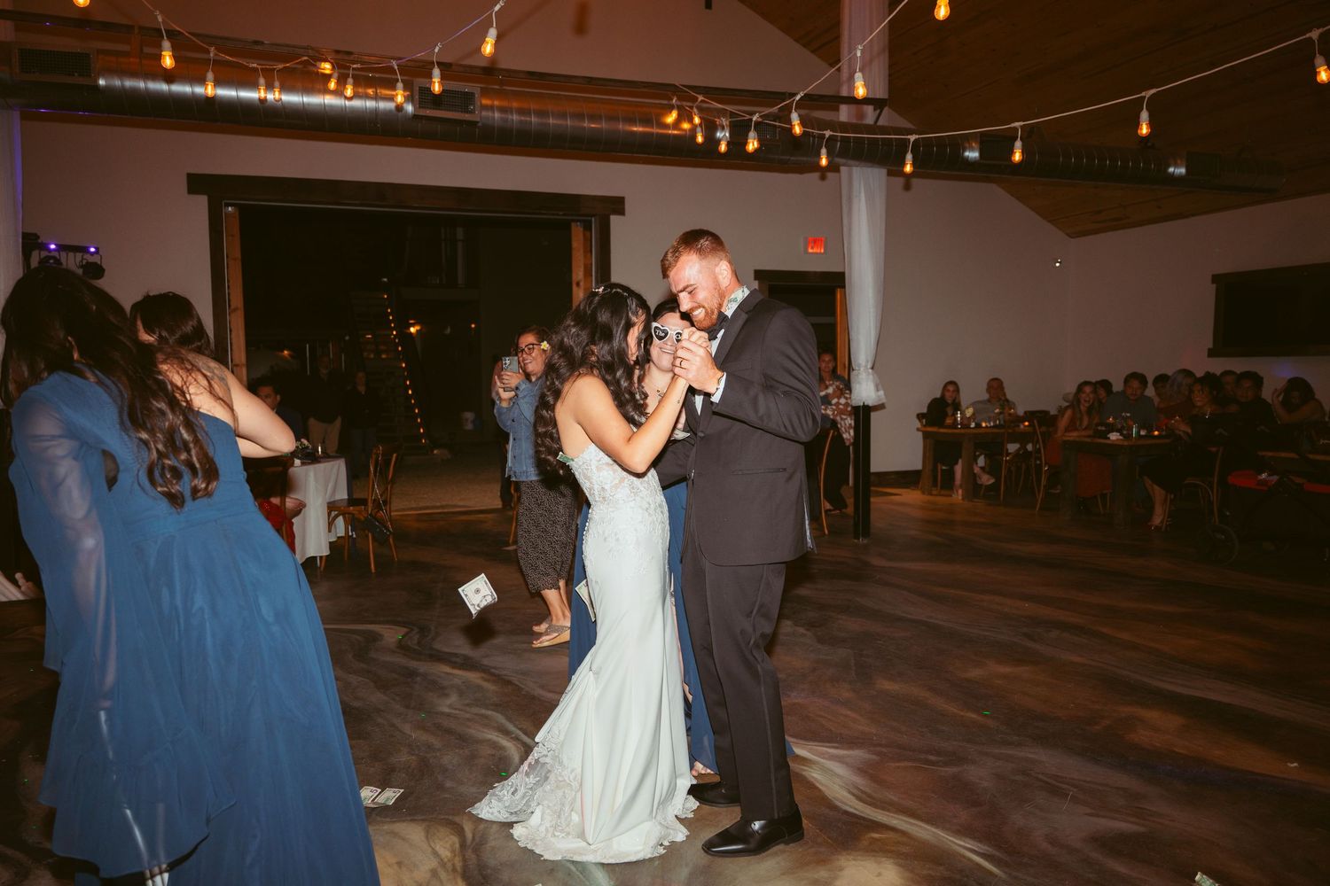 Wedding party dancing and celebrating in a rustic venue with string lighting.