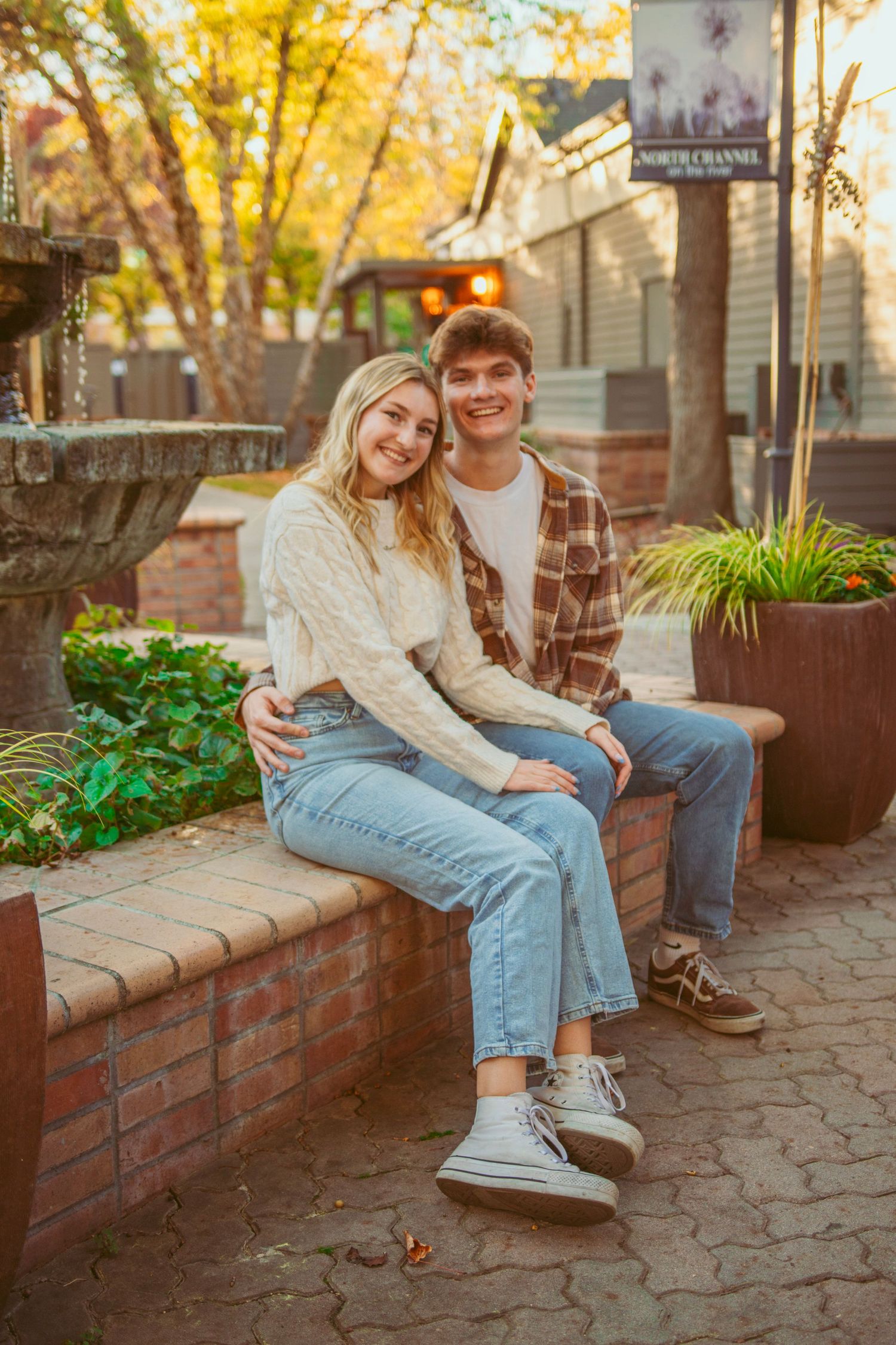 A couple relaxes on a brick wall in casual autumn attire.