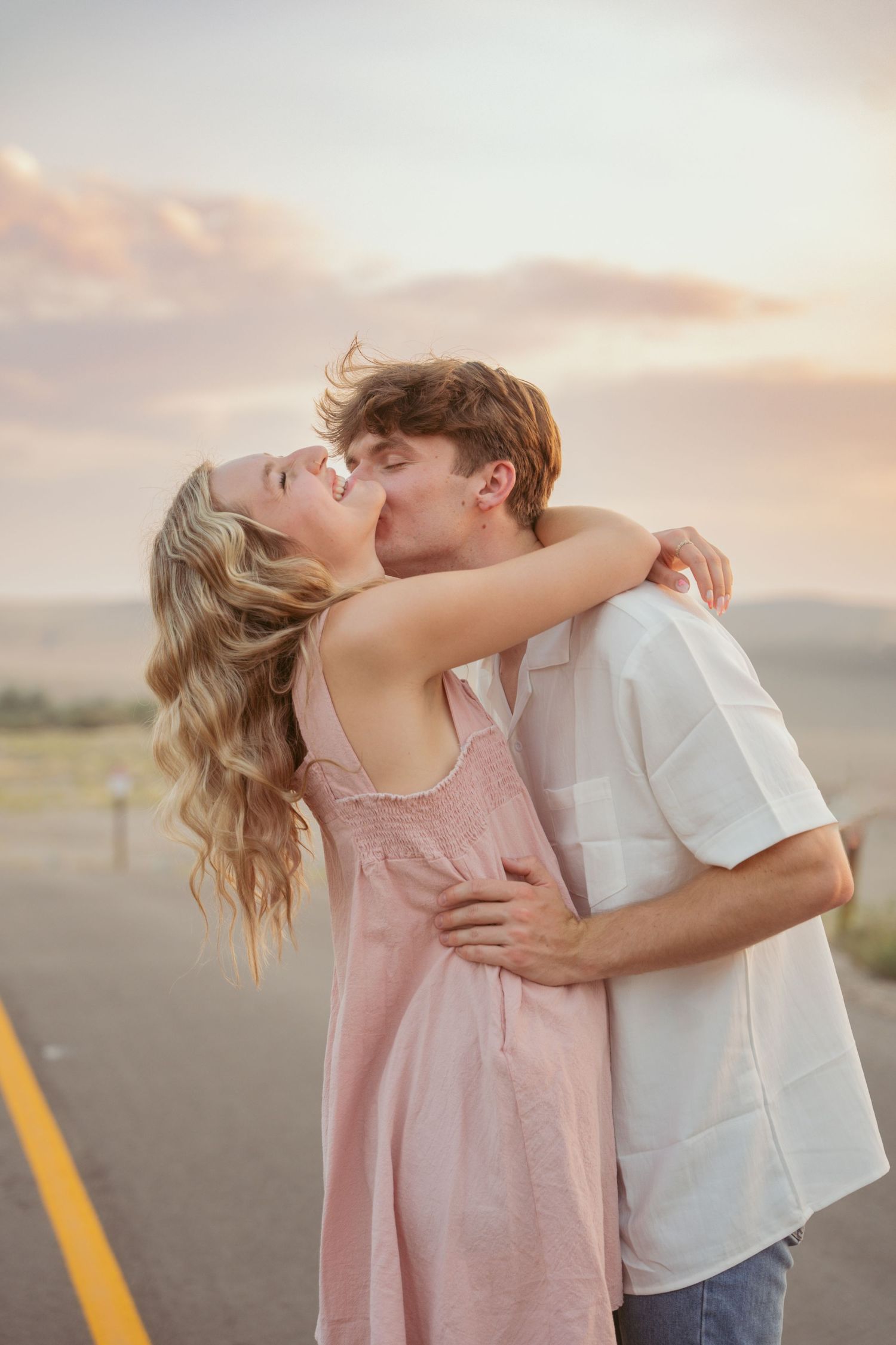 A couple in casual summer clothes shares a passionate kiss on a desert road at sunset.