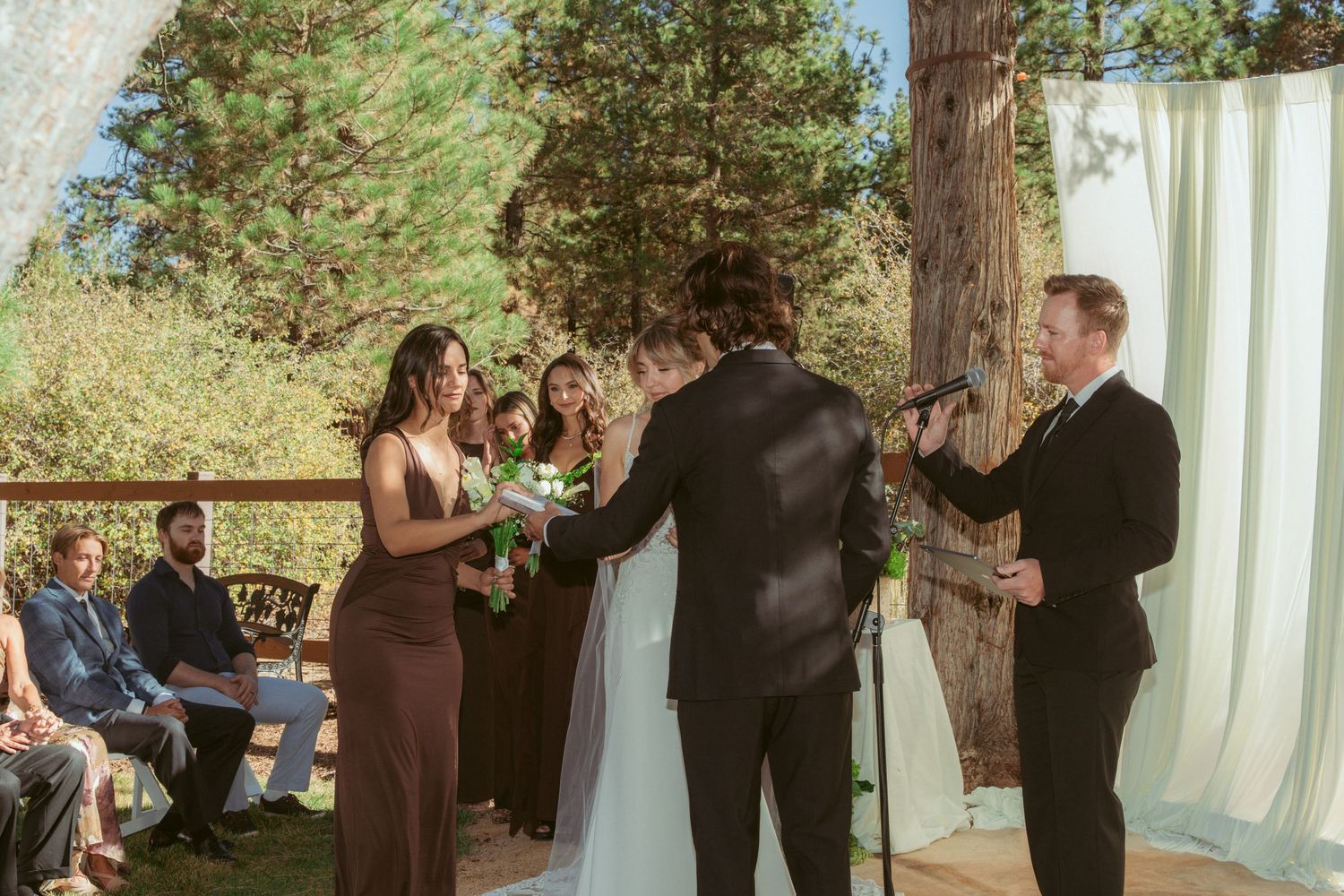 Wedding ceremony in sunlit forest with white drapes and brown-dressed attendants.