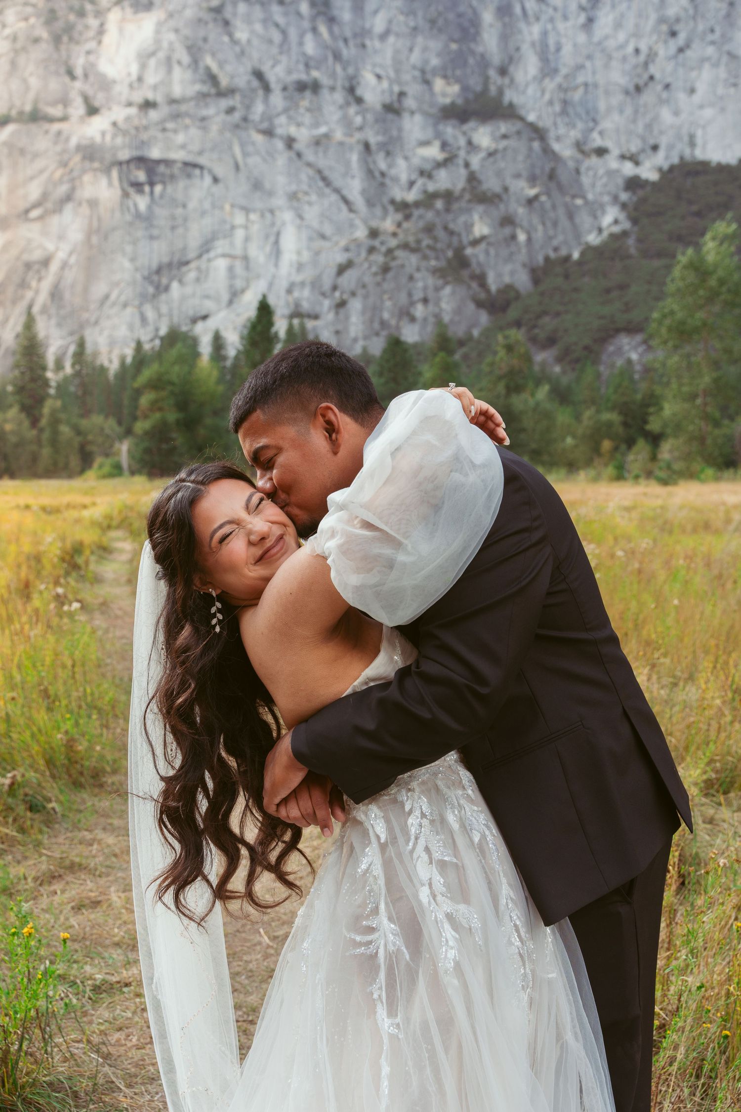 Wedding couple shares an intimate moment in a meadow with Yosemite's iconic cliffs behind them.