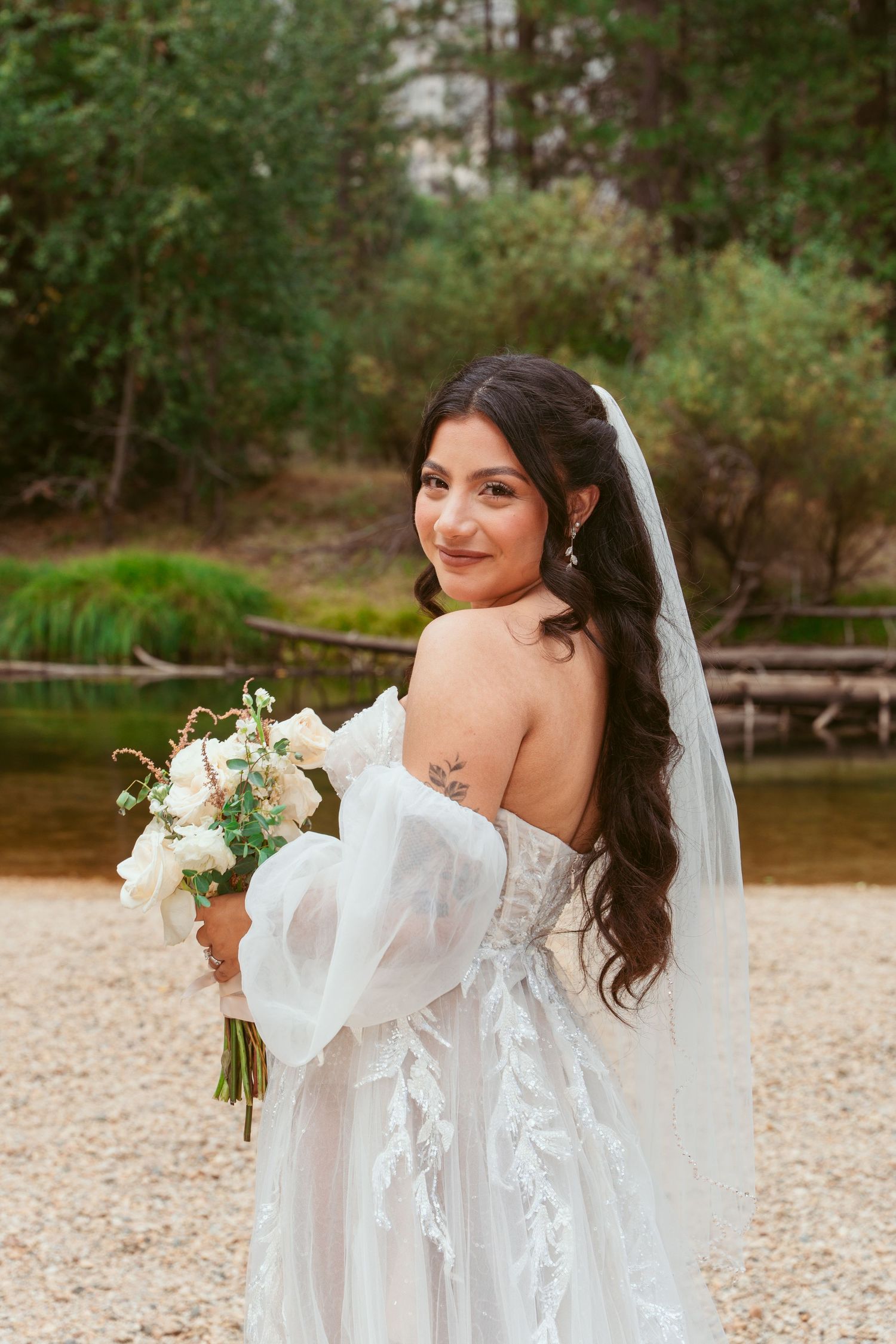 Bride in lace gown holding white bouquet against mountain landscape backdrop.