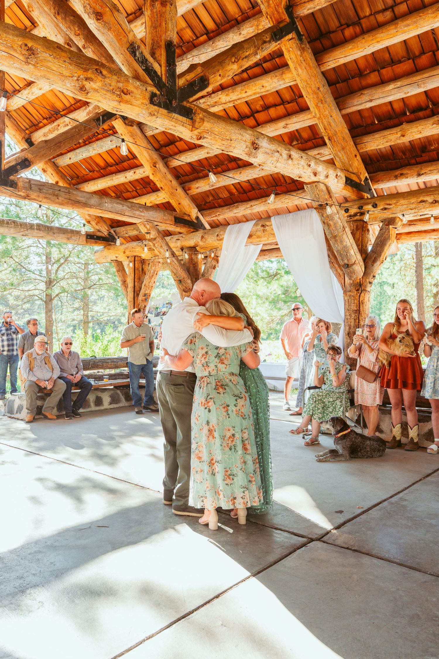 Wedding guests dance under a rustic wooden pavilion decorated with white curtains and string lights.
