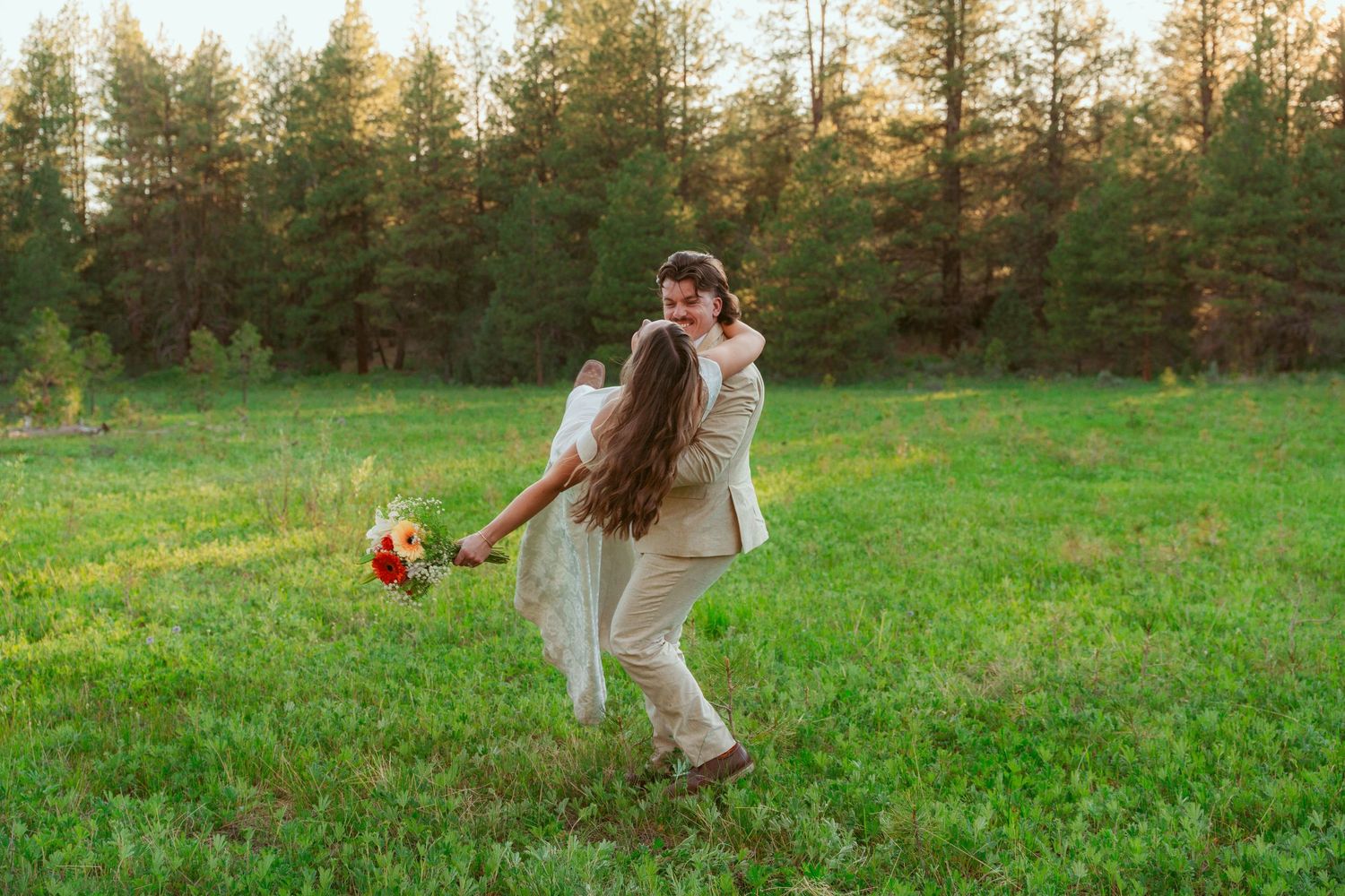 Couple running playfully through a green field at sunset with pine trees in background.