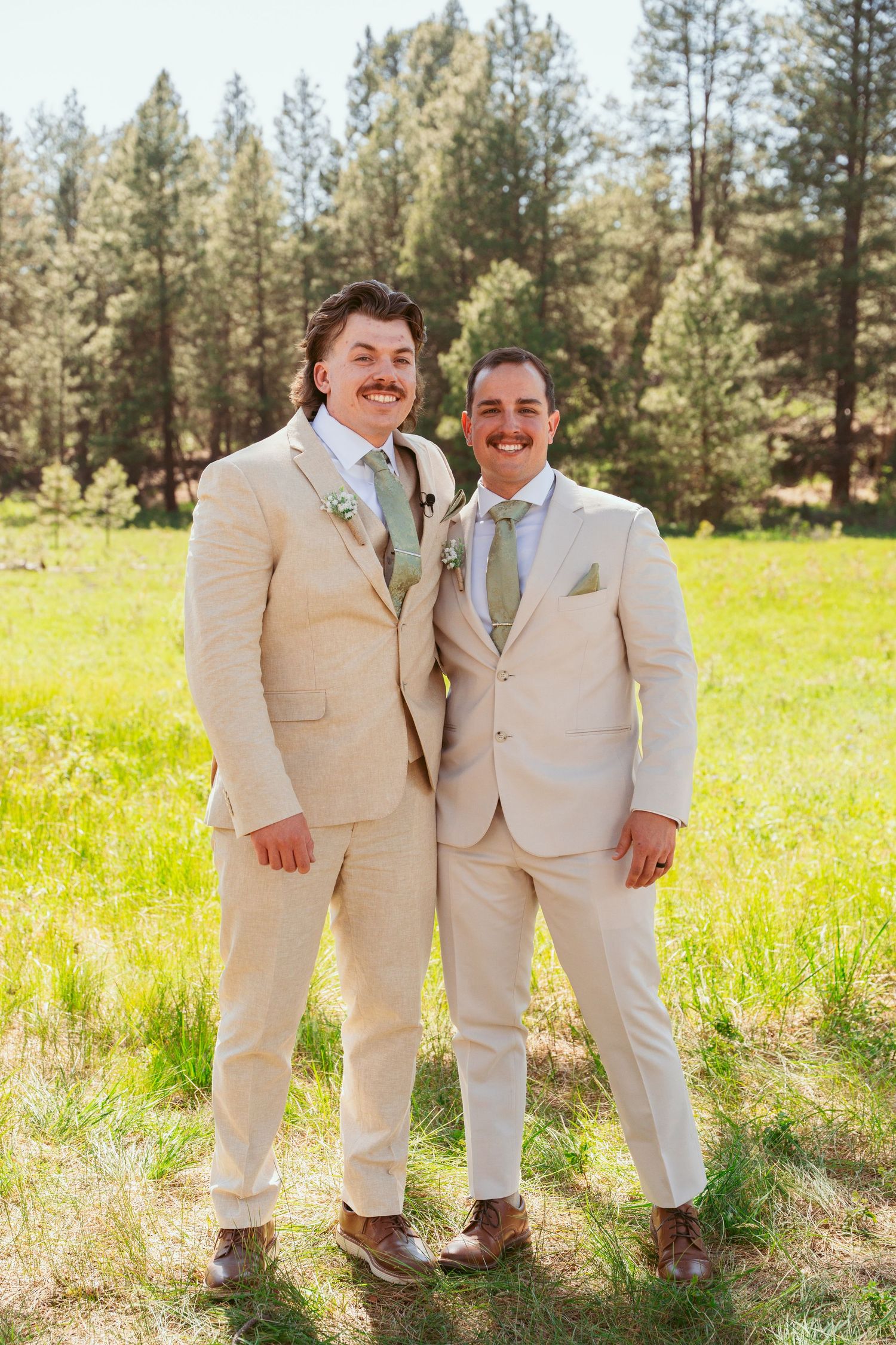 Two people in matching light suits share a joyful moment in a meadow with evergreens.