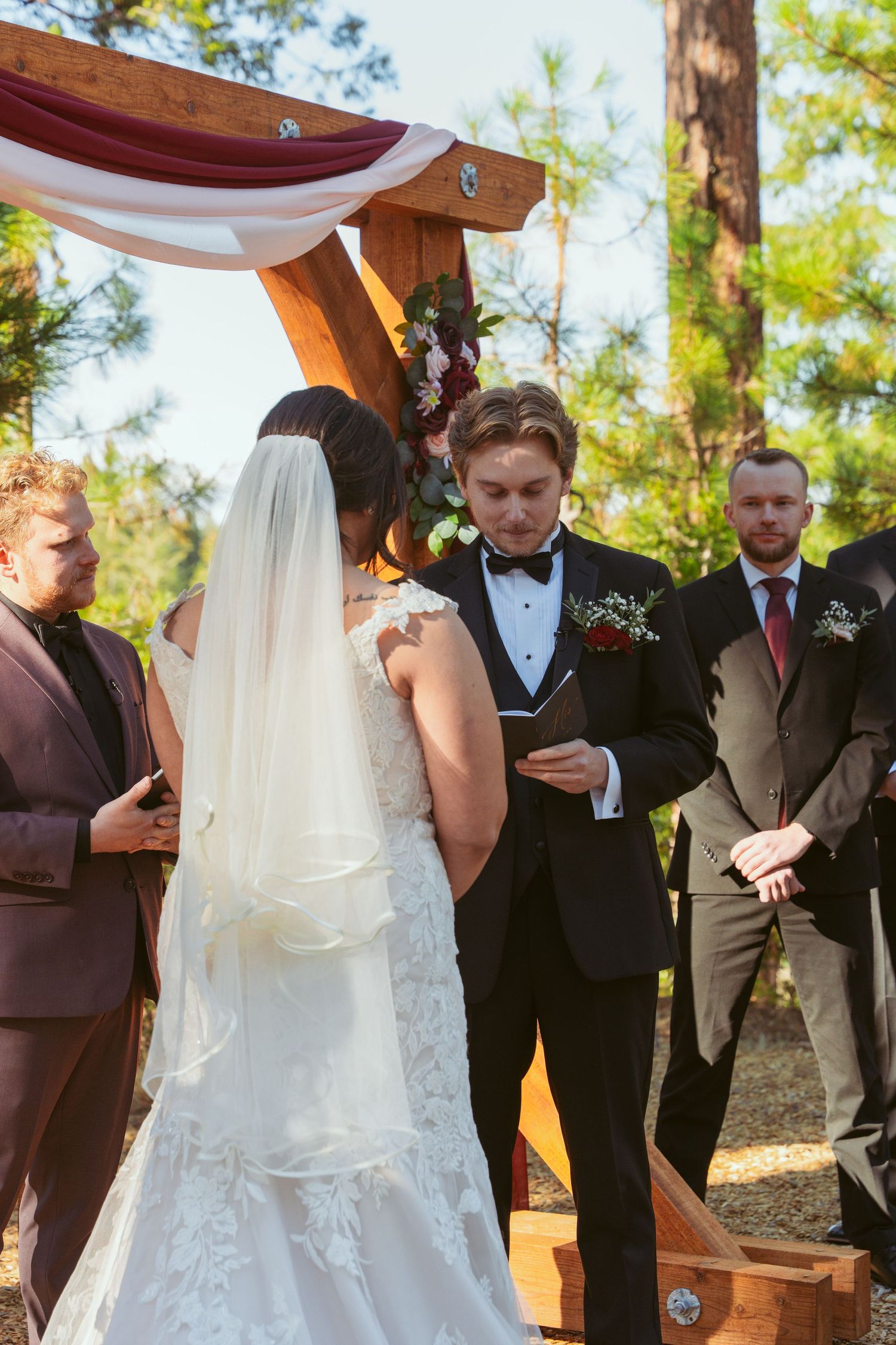 Wedding ceremony participants gather under wooden arch with white and burgundy draping.