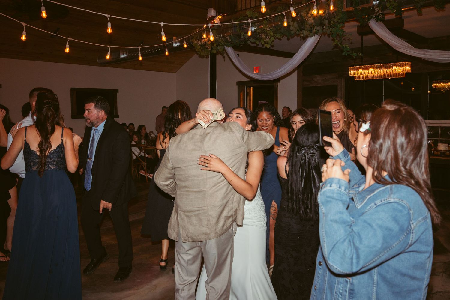Wedding guests dancing under string lights at indoor reception venue.