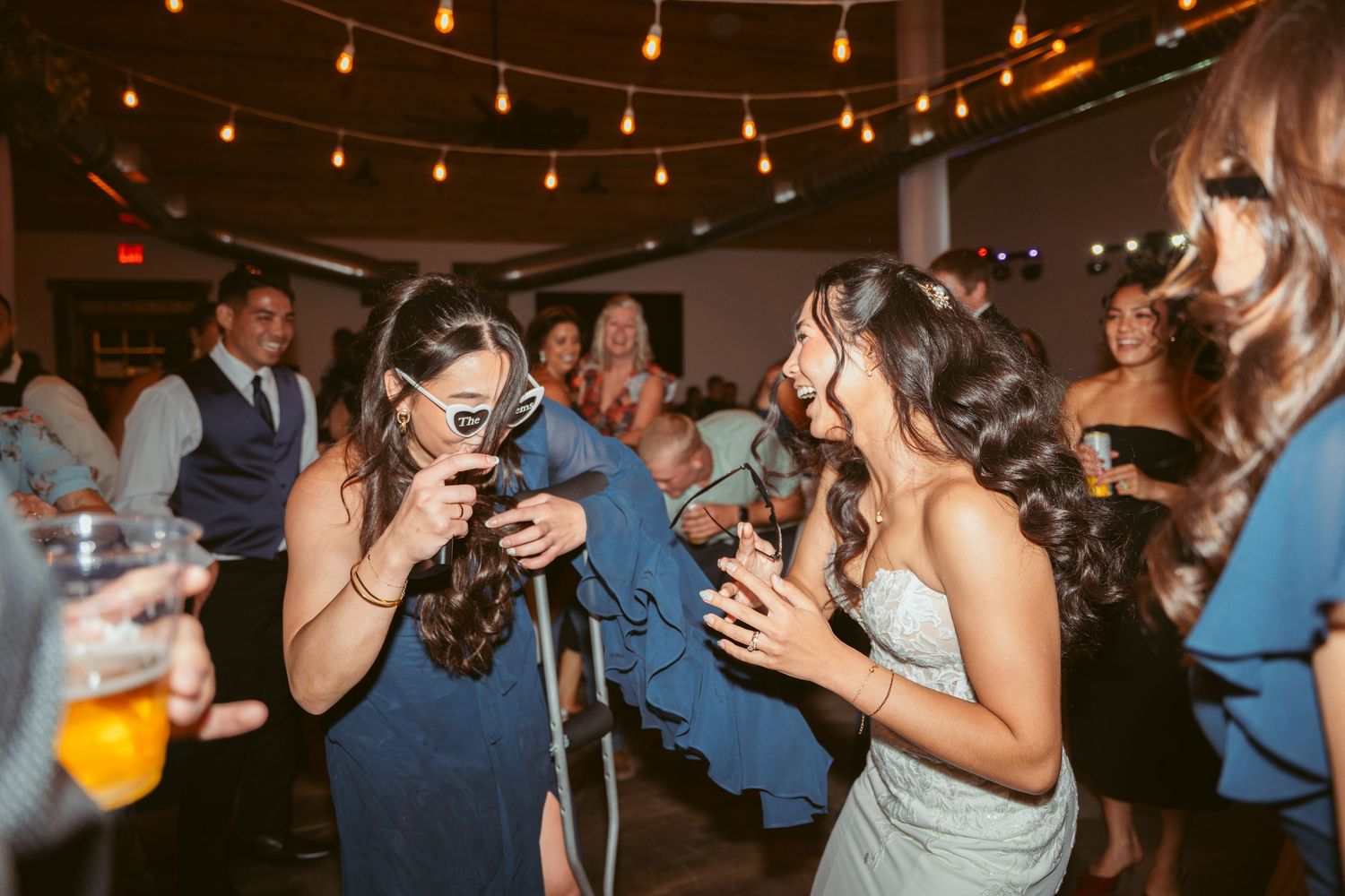 Guests in formal attire laugh and dance under string lights at indoor celebration.