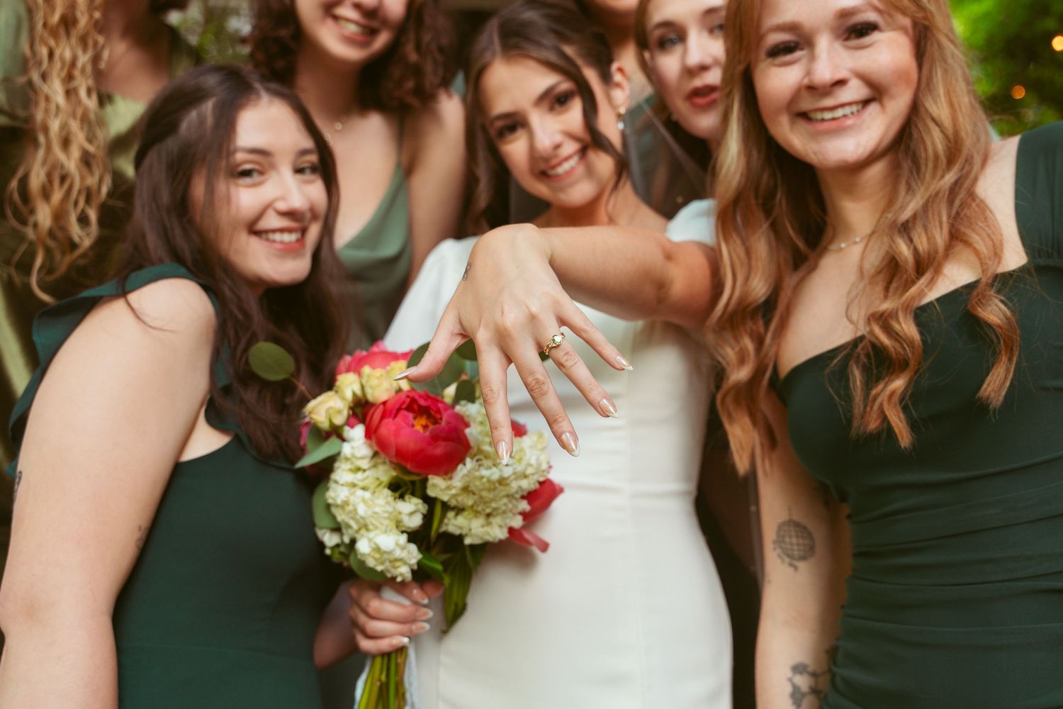 Wedding party members in dark green dresses and white gown pose together on red porch with smiles and flowers.