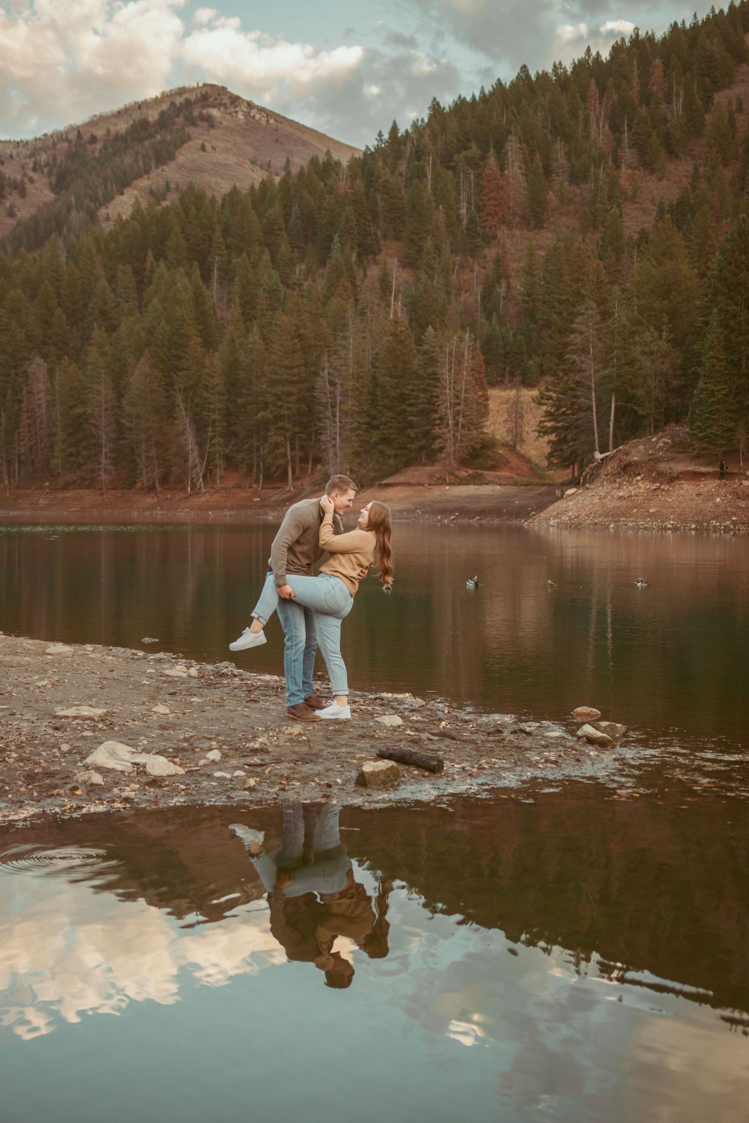 Romantic couple poses together by a serene mountain lake at sunset with reflections in the calm water.