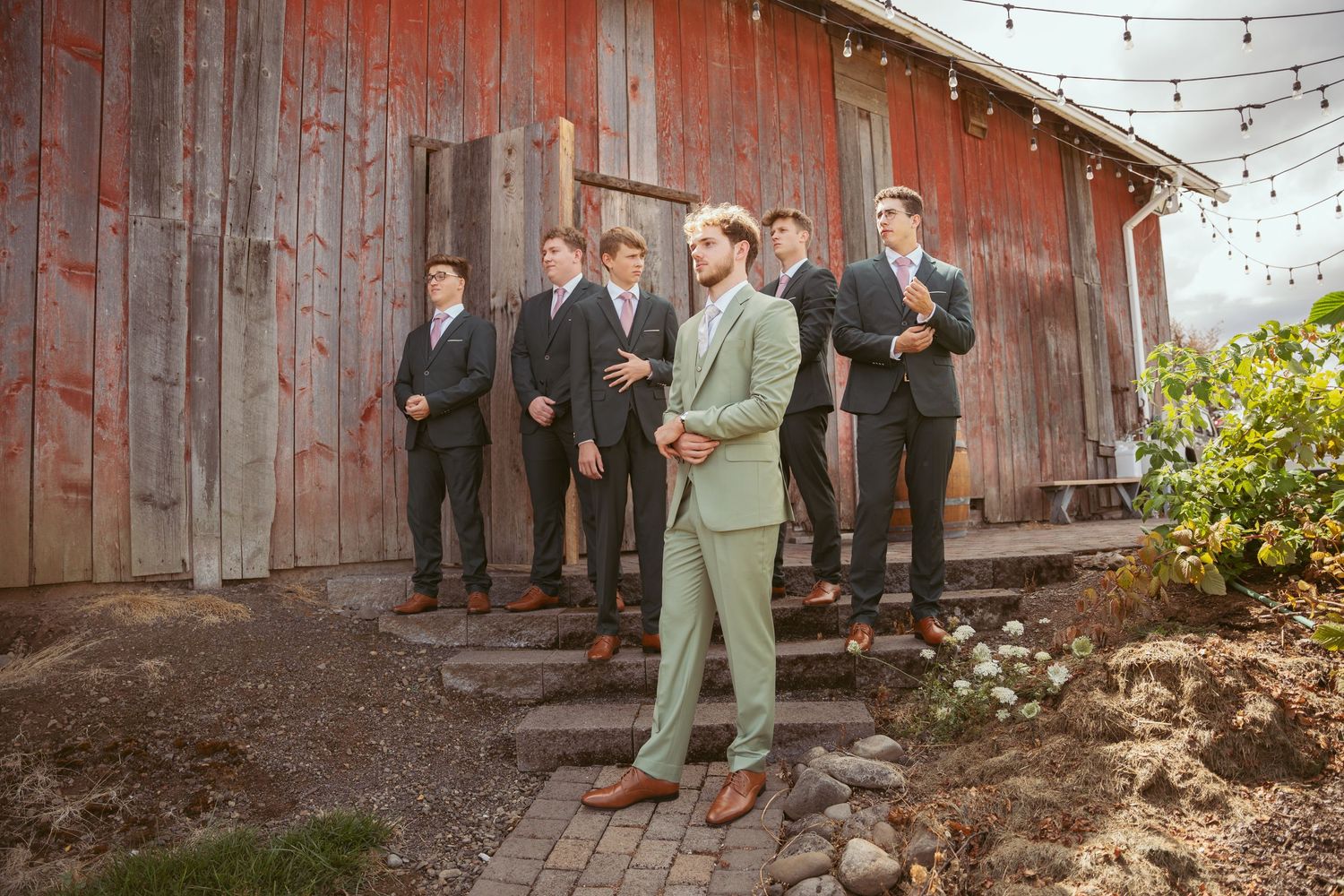 Wedding party members stand in formation by weathered red barn with one wearing sage green suit.