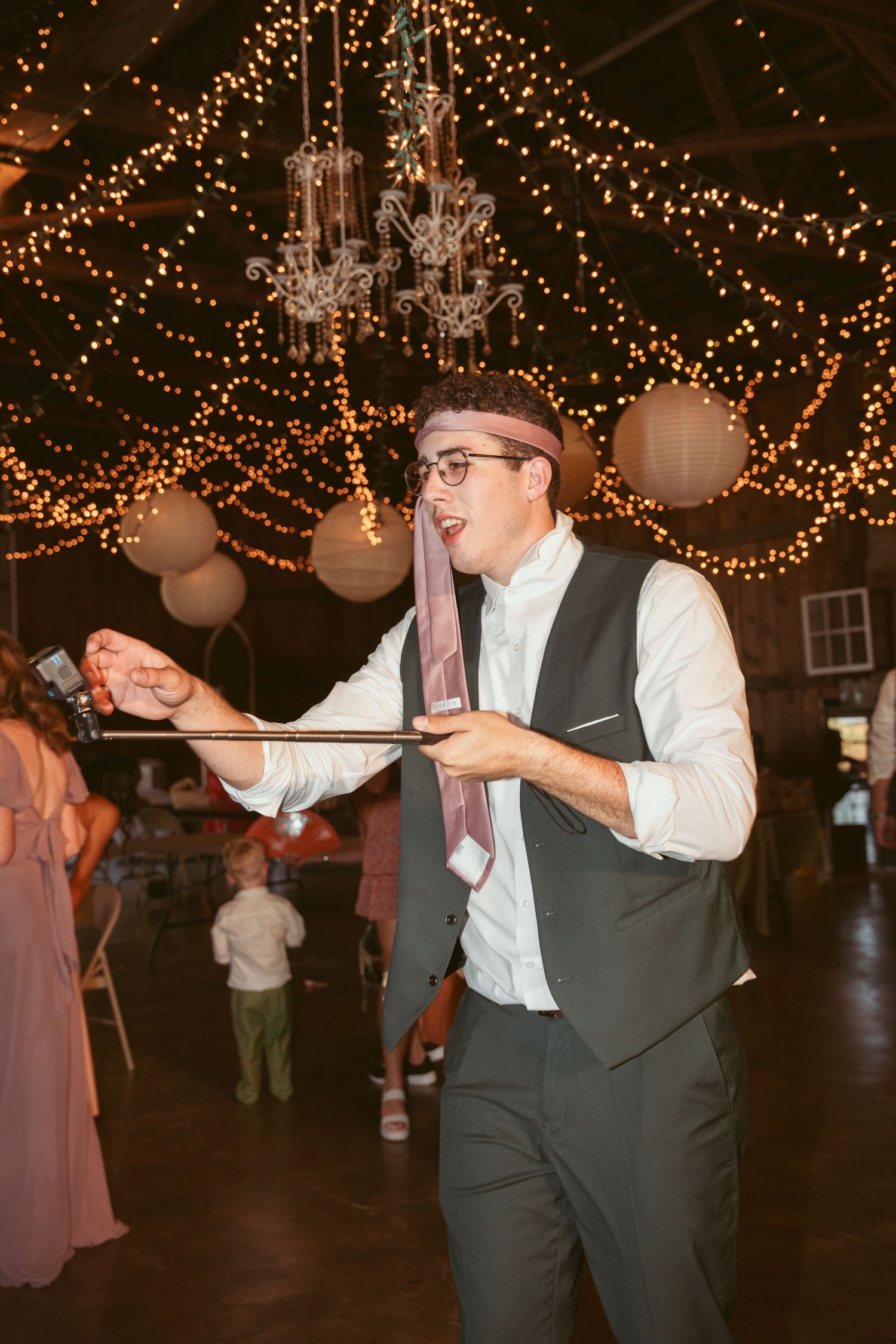 Wedding guests dancing under string lights and white lanterns in a rustic barn setting.