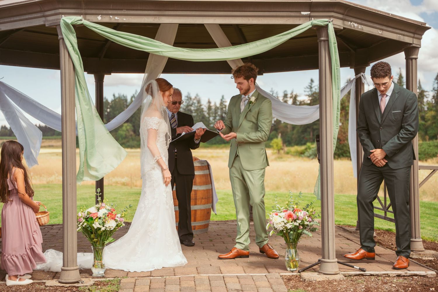 Wedding ceremony participants standing under decorated wooden pavilion in countryside setting.