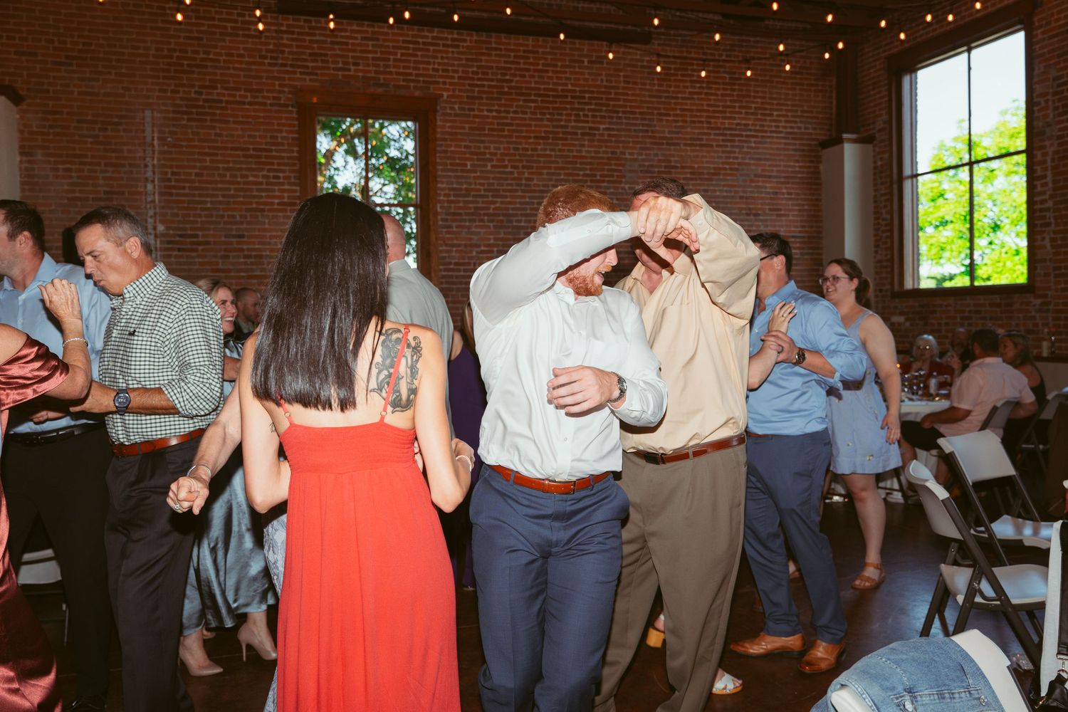 Guests dancing and socializing at a wedding reception in an industrial-style venue with string lights.