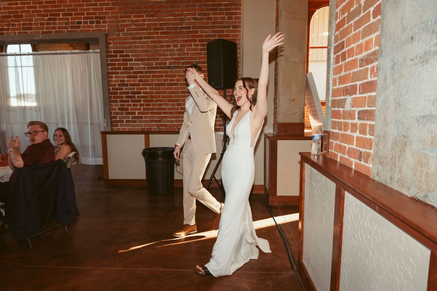 Wedding party dancing and celebrating in a rustic brick venue with natural lighting.