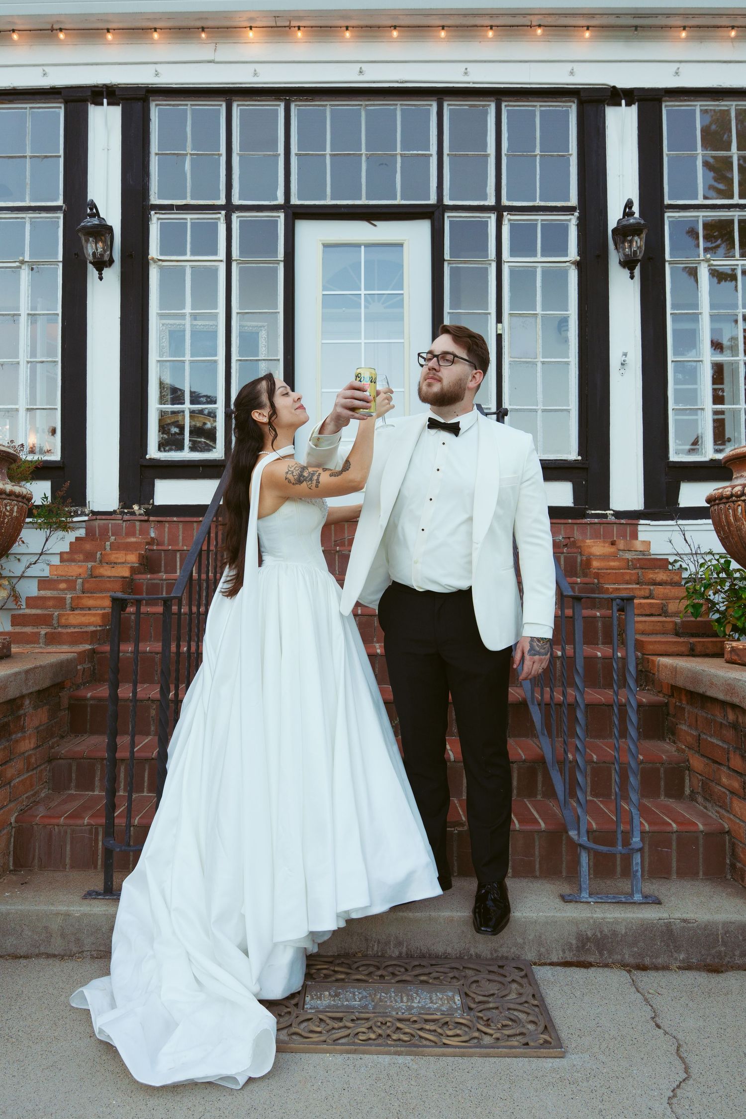 A newlywed couple toasts with champagne glasses in front of a venue with large windows at dusk.