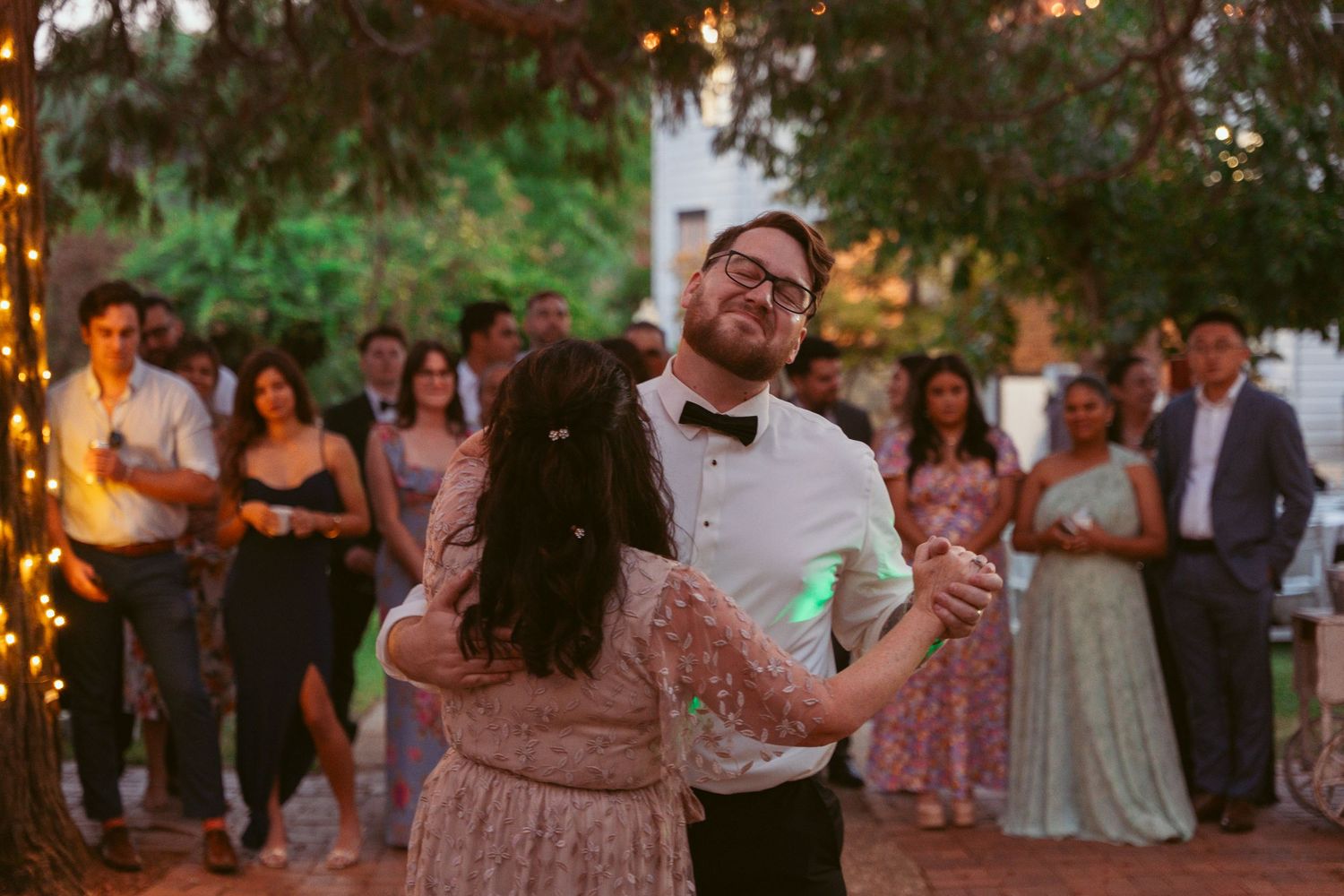 Outdoor evening wedding reception with string lights and dancing guests.