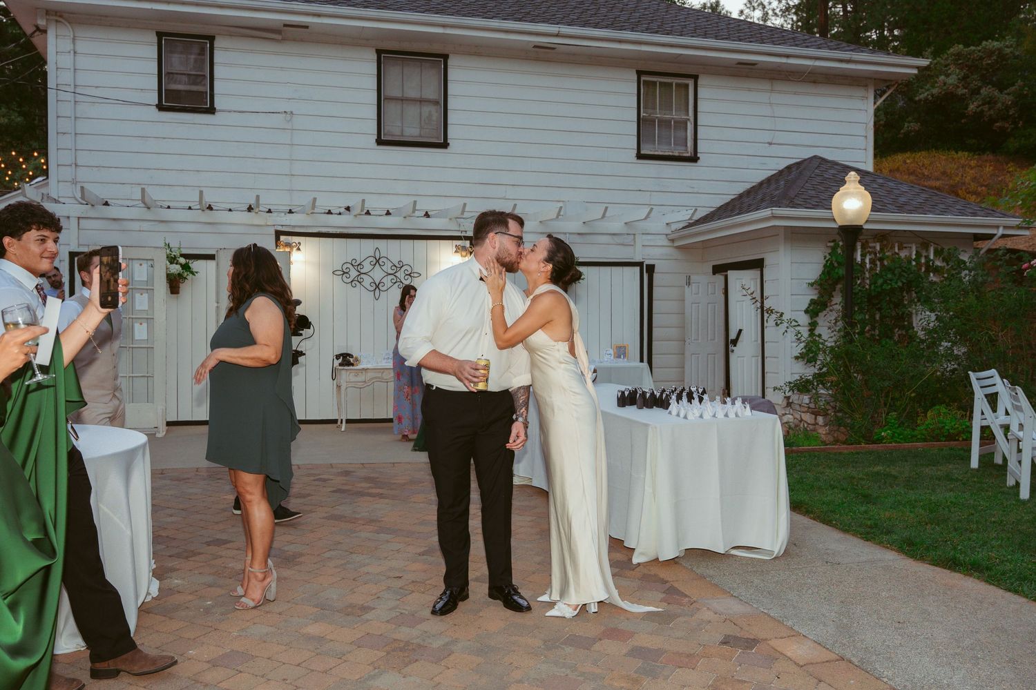 Outdoor wedding reception at a white house with string lights and guests mingling on the patio.
