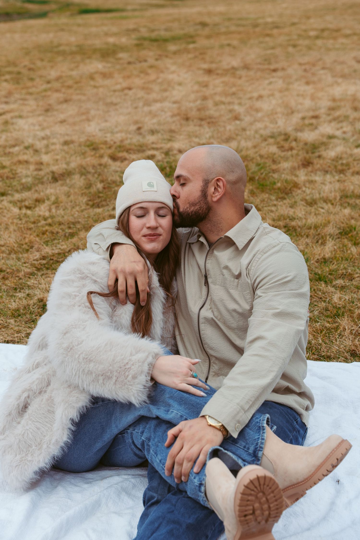 Couple in casual winter wear sharing intimate moments while sitting on white blanket outdoors.