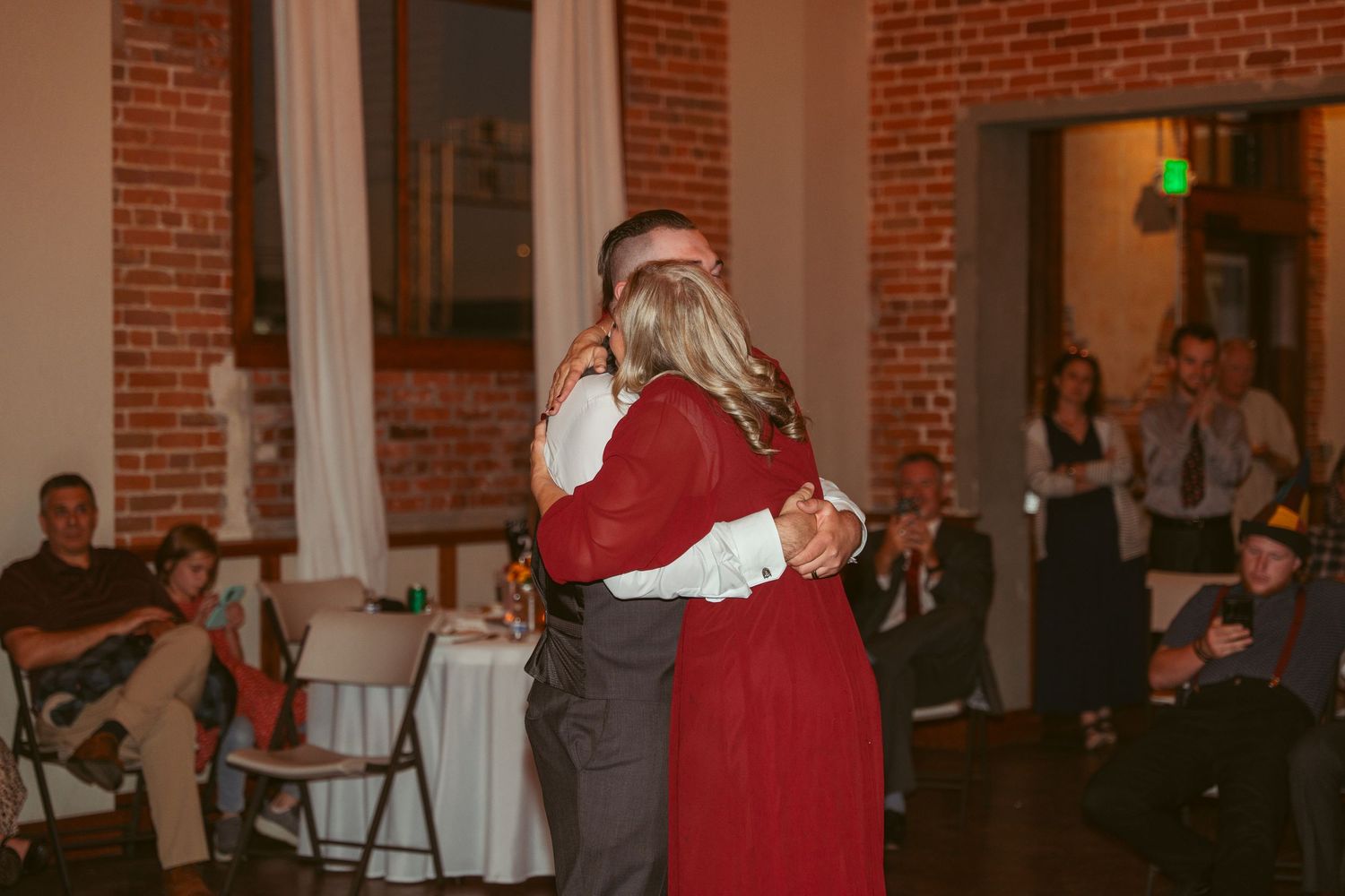 Wedding guests dance and celebrate on a dance floor in a rustic brick venue with string lights overhead.