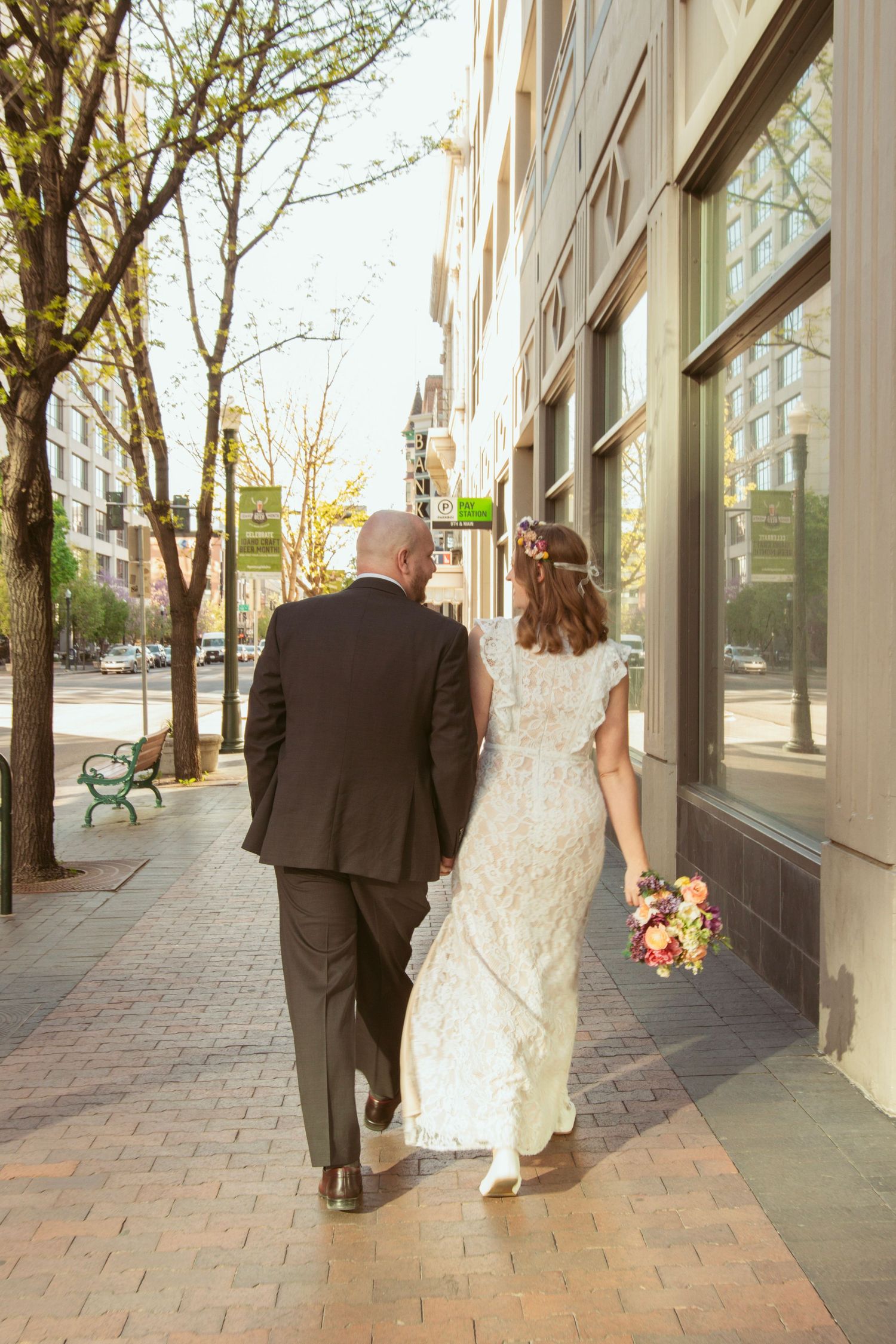 A couple walks down an urban sidewalk with spring trees and storefronts.