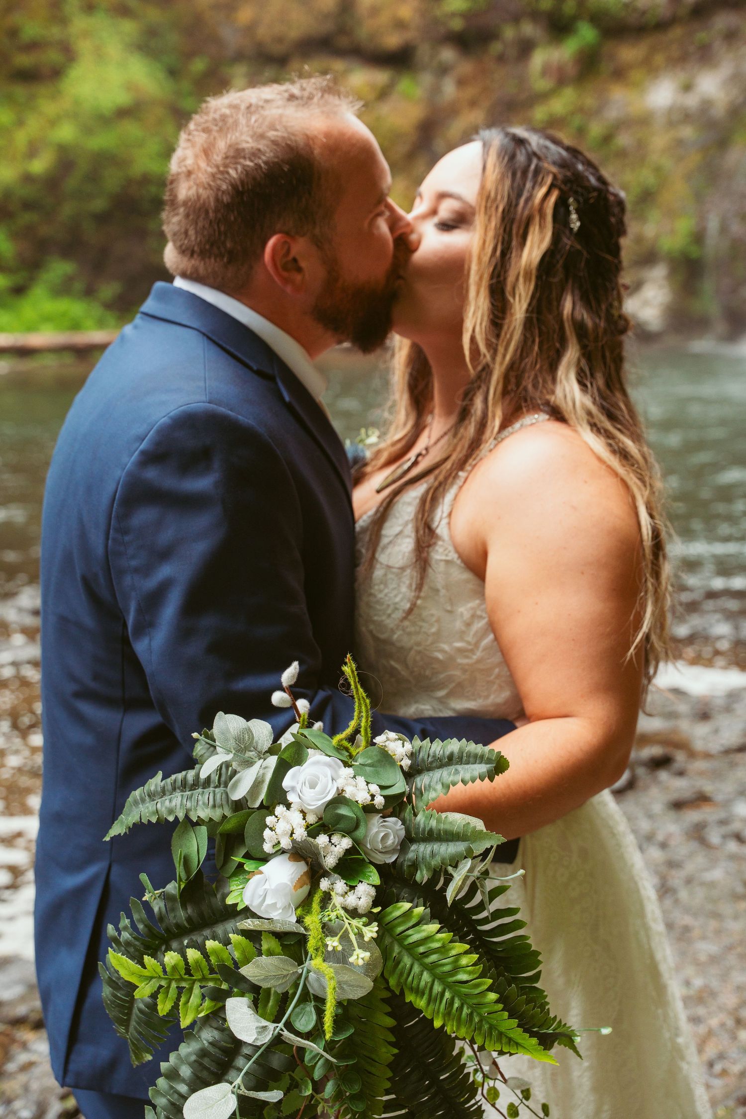 Intimate wedding portrait with fern bouquet against natural waterfall backdrop.